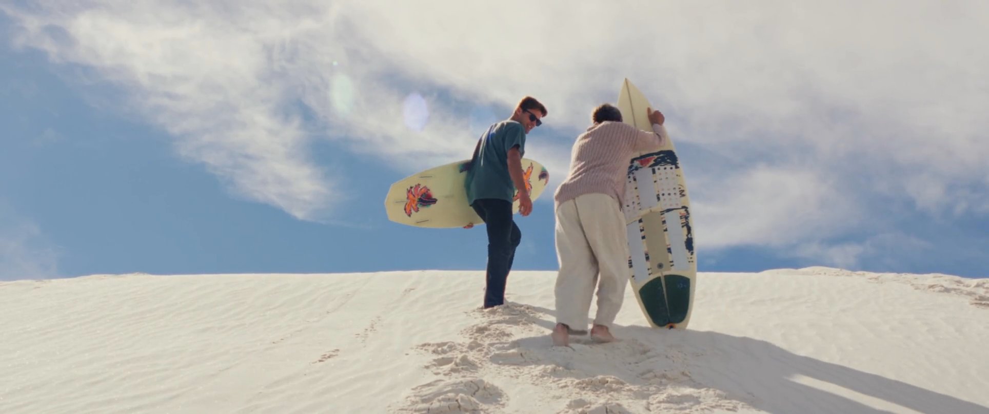 Two individuals carry surfboards while traversing a steep desert sand dune under a bright, sunny sky.