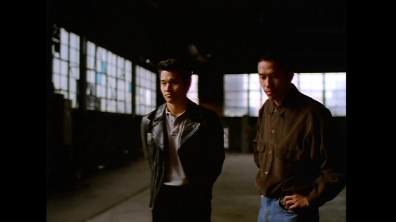 Two men discuss a potential transaction in a dimly lit, expansive industrial warehouse, centered around a silver metal briefcase.