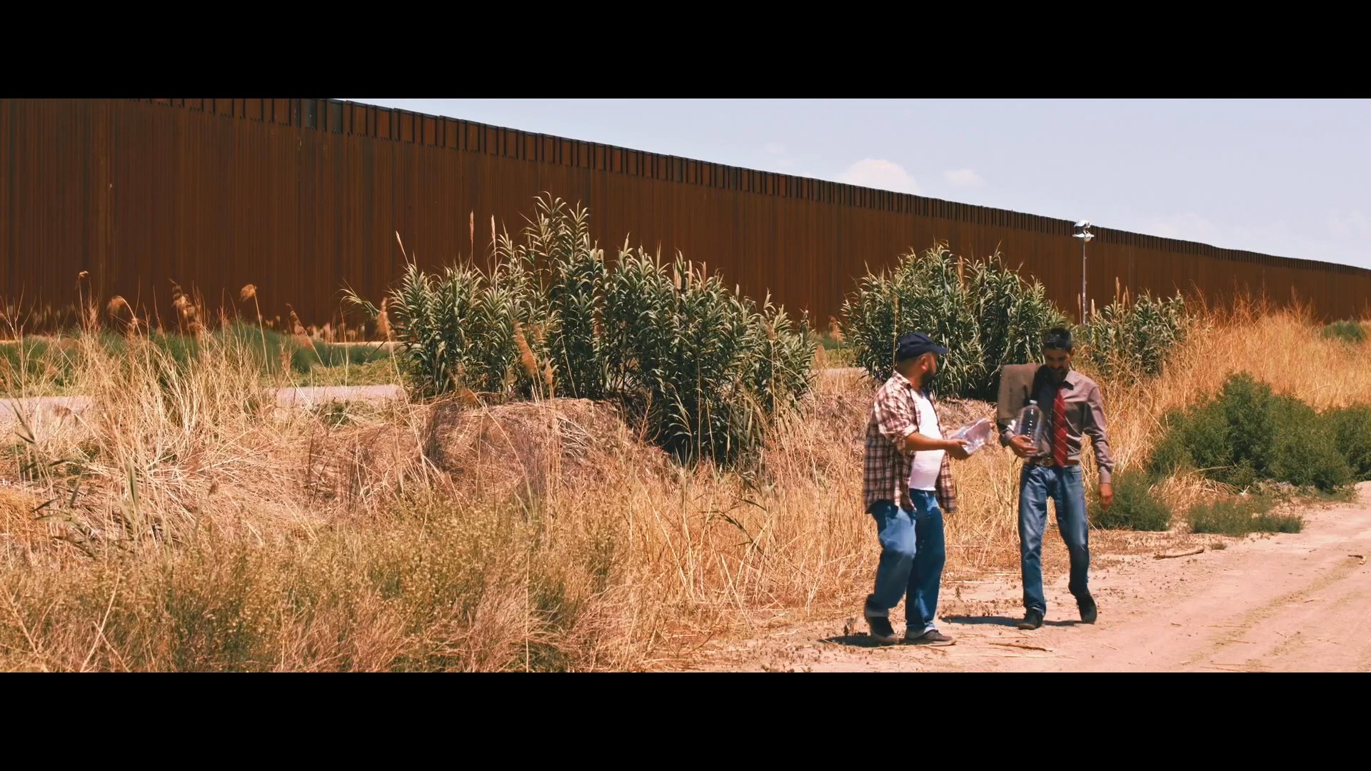 A man expresses frustration toward a border wall while his companions attempt to navigate the arid terrain. The group discusses the logistical challenges of transporting their belongings across the landscape.