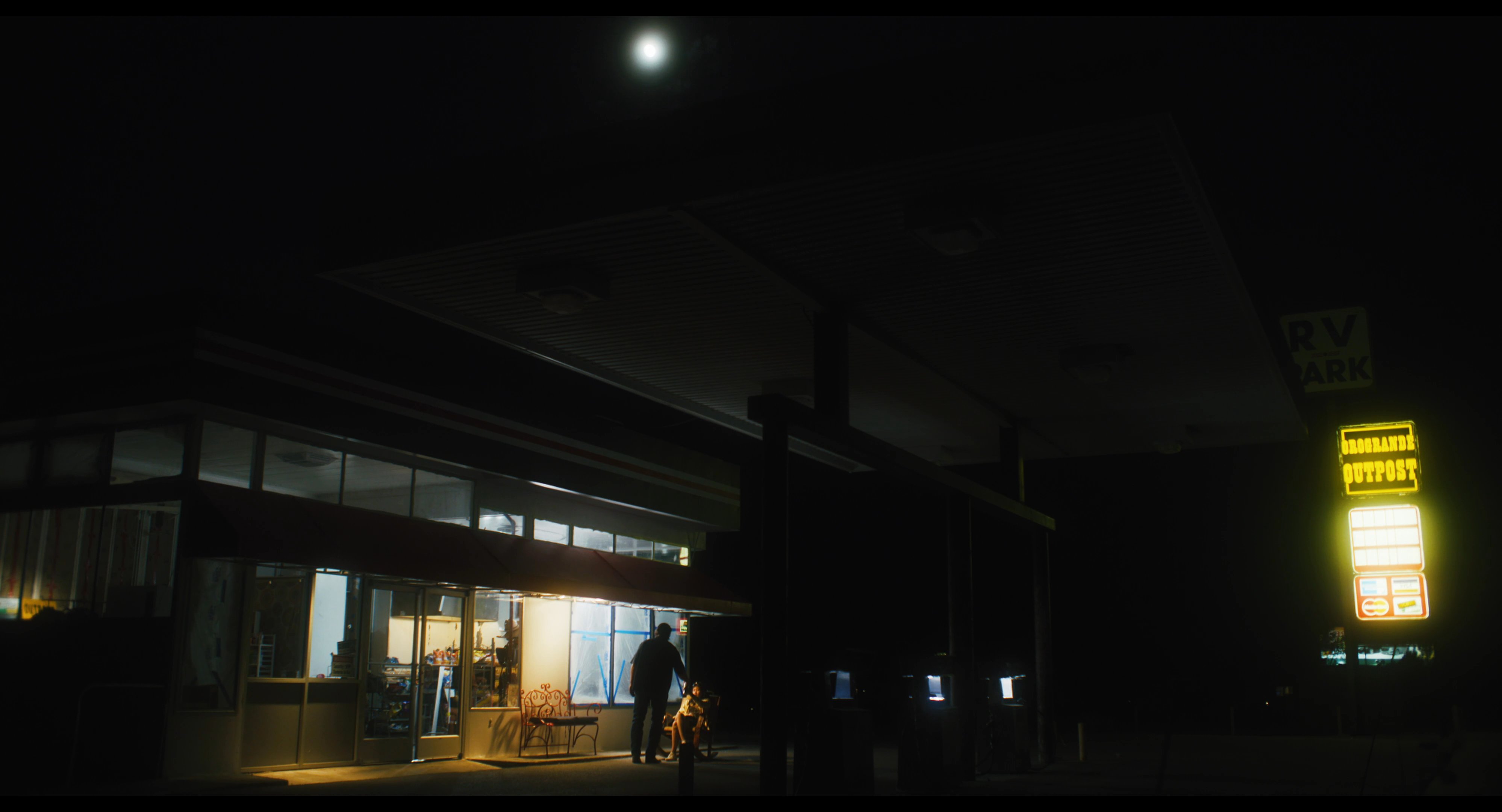 A man and a woman engage in conversation while sitting outside a brightly lit gas station at night. The pair rests on a bench and a rocking chair, illuminated by the establishment's store front lights.