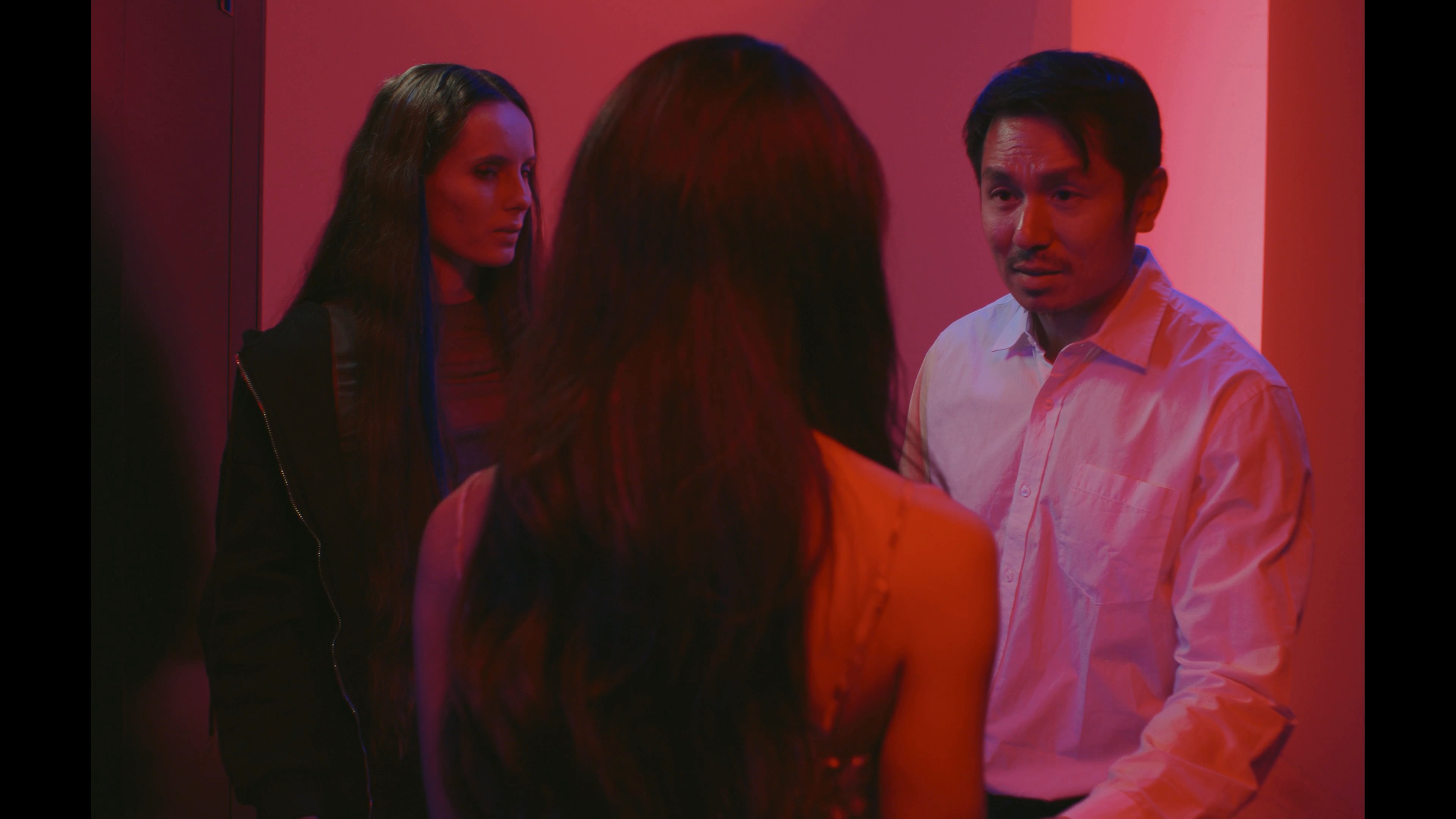 Three individuals engage in a tense conversation within a brightly lit red space. The interaction intensifies as a man and woman share an intimate moment while a third person observes.