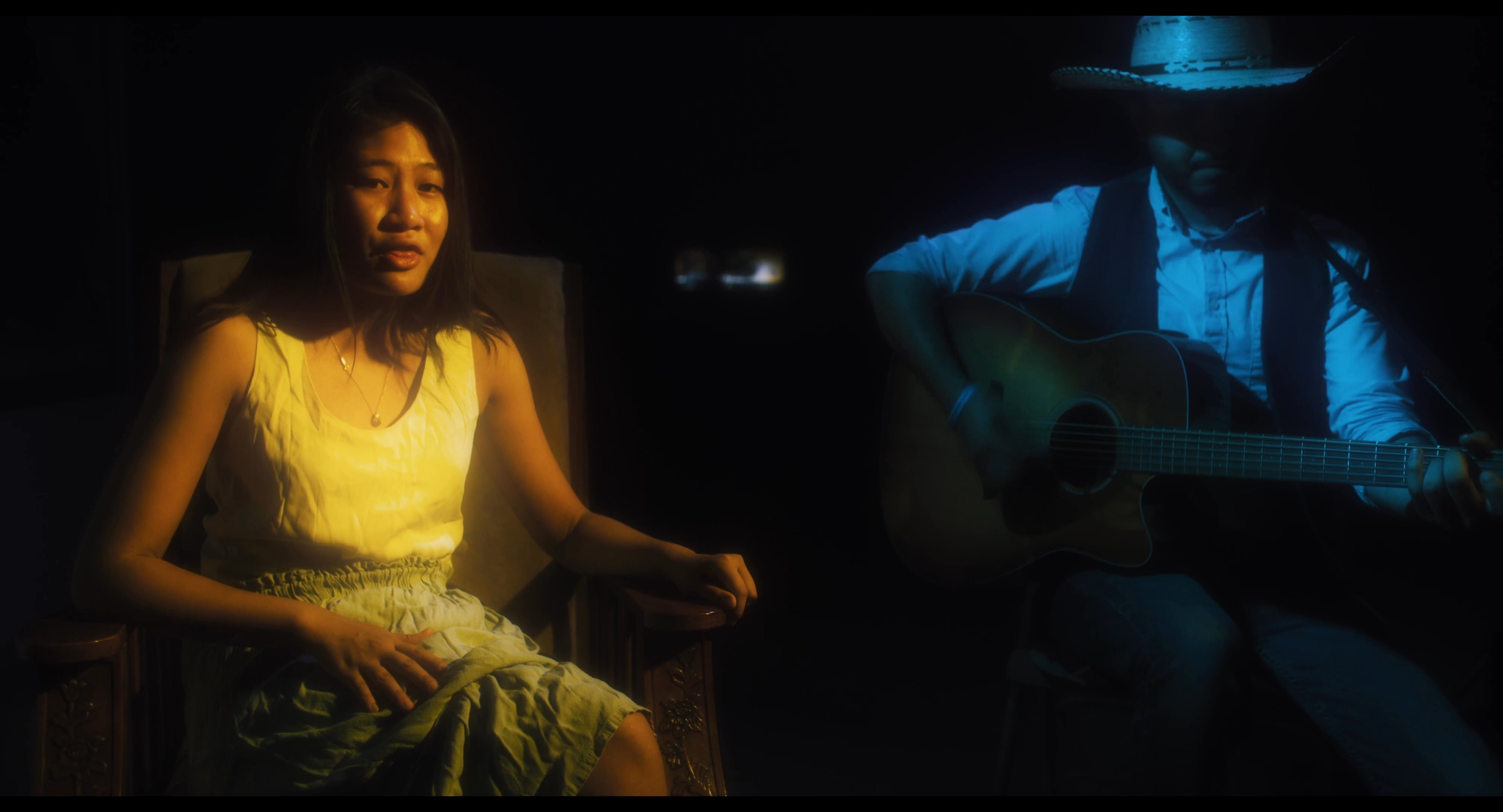 A female singer performs an acoustic song alongside a guitarist in a dimly lit studio setting.