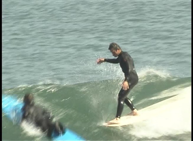 A surfer in a black wetsuit skillfully maneuvers along the face of a rolling ocean wave. The individual maintains balance and control while gliding across the water's surface.
