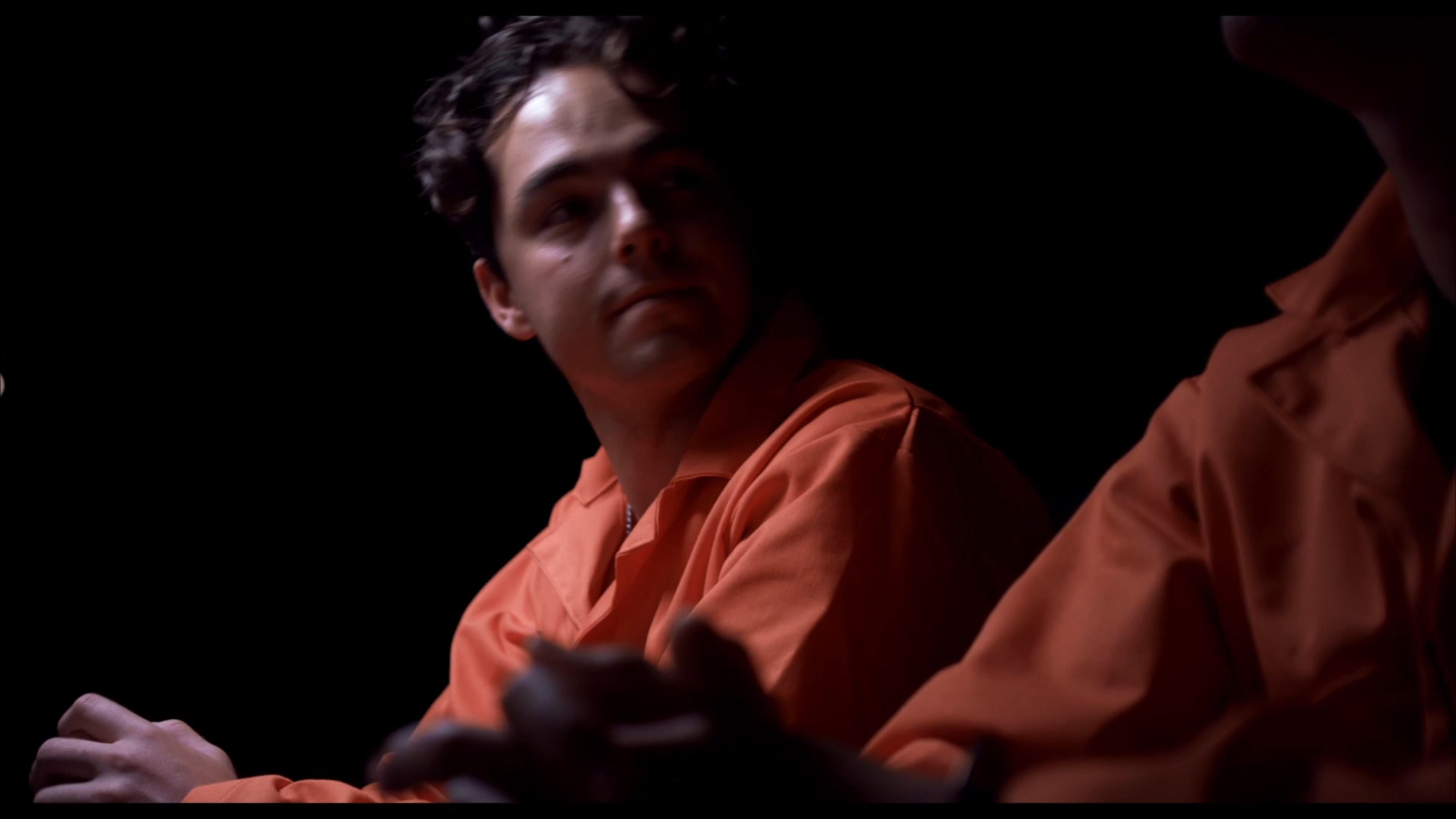 Two individuals wearing orange prison jumpsuits and handcuffs sit in a dimly lit room, engaged in a tense conversation. The setting suggests an interrogation or legal briefing environment.