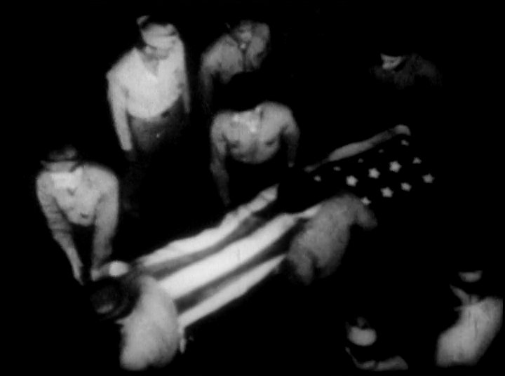 United States military personnel conduct a solemn burial at sea ceremony with a flag-draped casket on a ship deck. Servicemen gather in a disciplined formation to pay their final respects during the memorial.