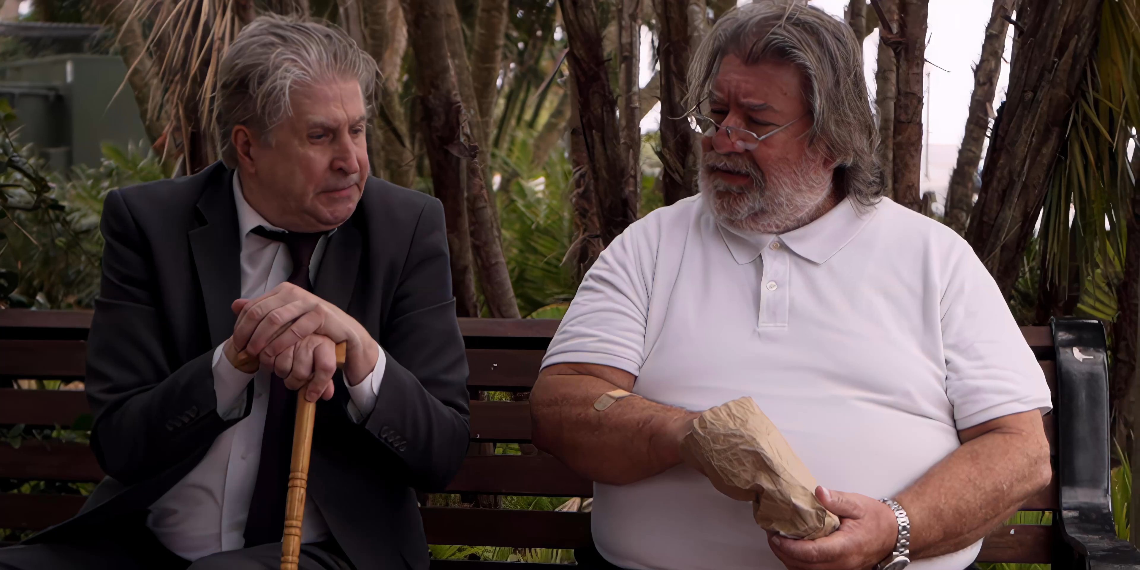 Two men sit on a park bench and engage in a seated conversation, with one man using a wooden cane.