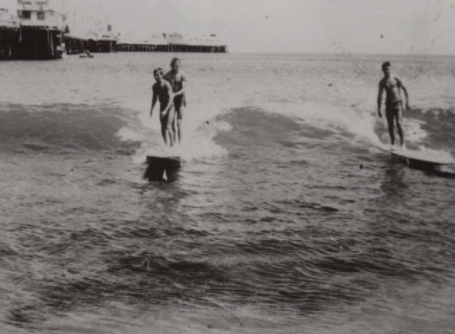 An elderly individual observes and discusses historical black-and-white film of people surfing. The footage provides a visual contrast between the narrator's present-day reflection and his documented personal history.