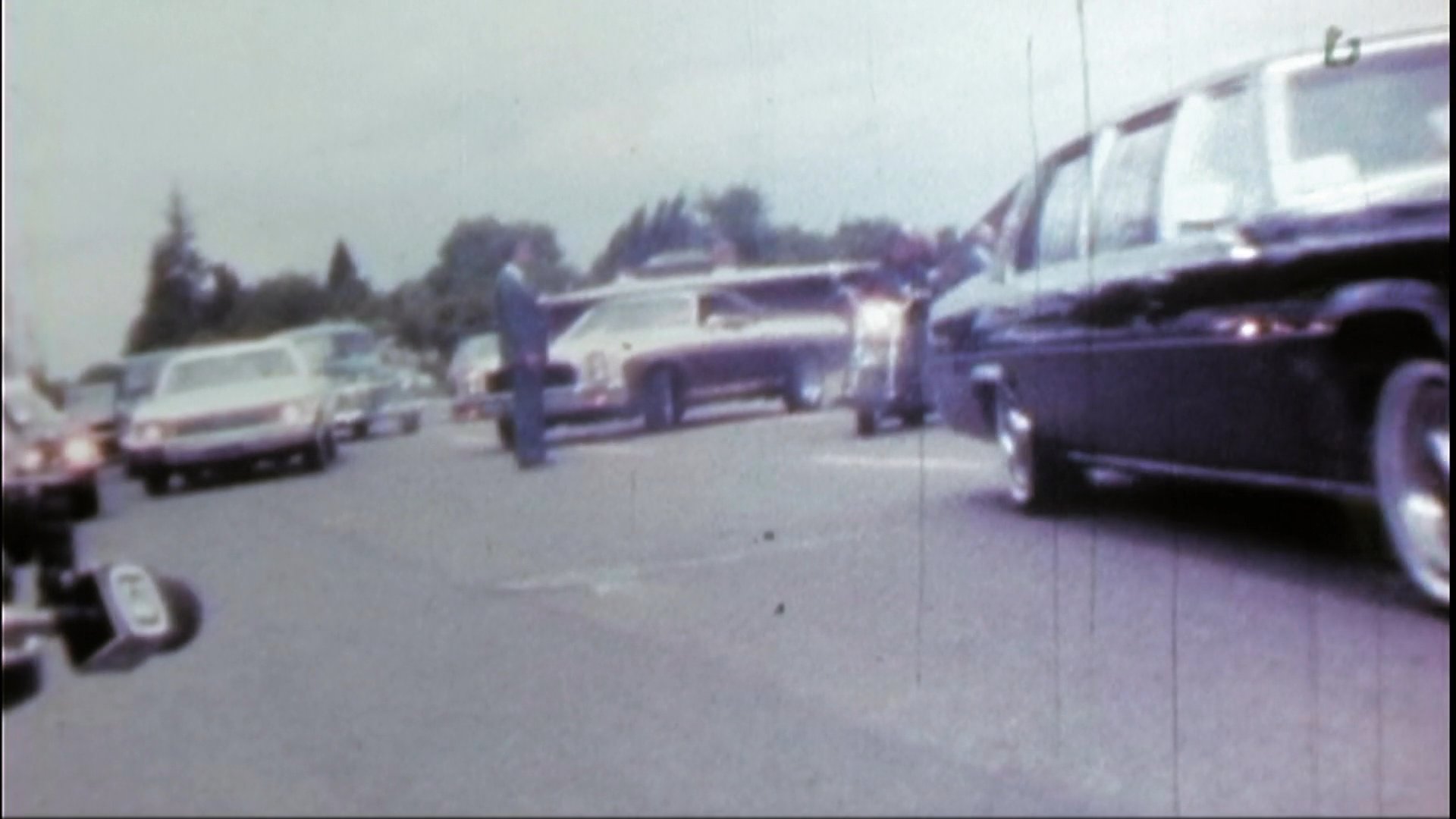 A group of motorcyclists participates in a somber funeral procession, following a hearse on an outdoor roadway.