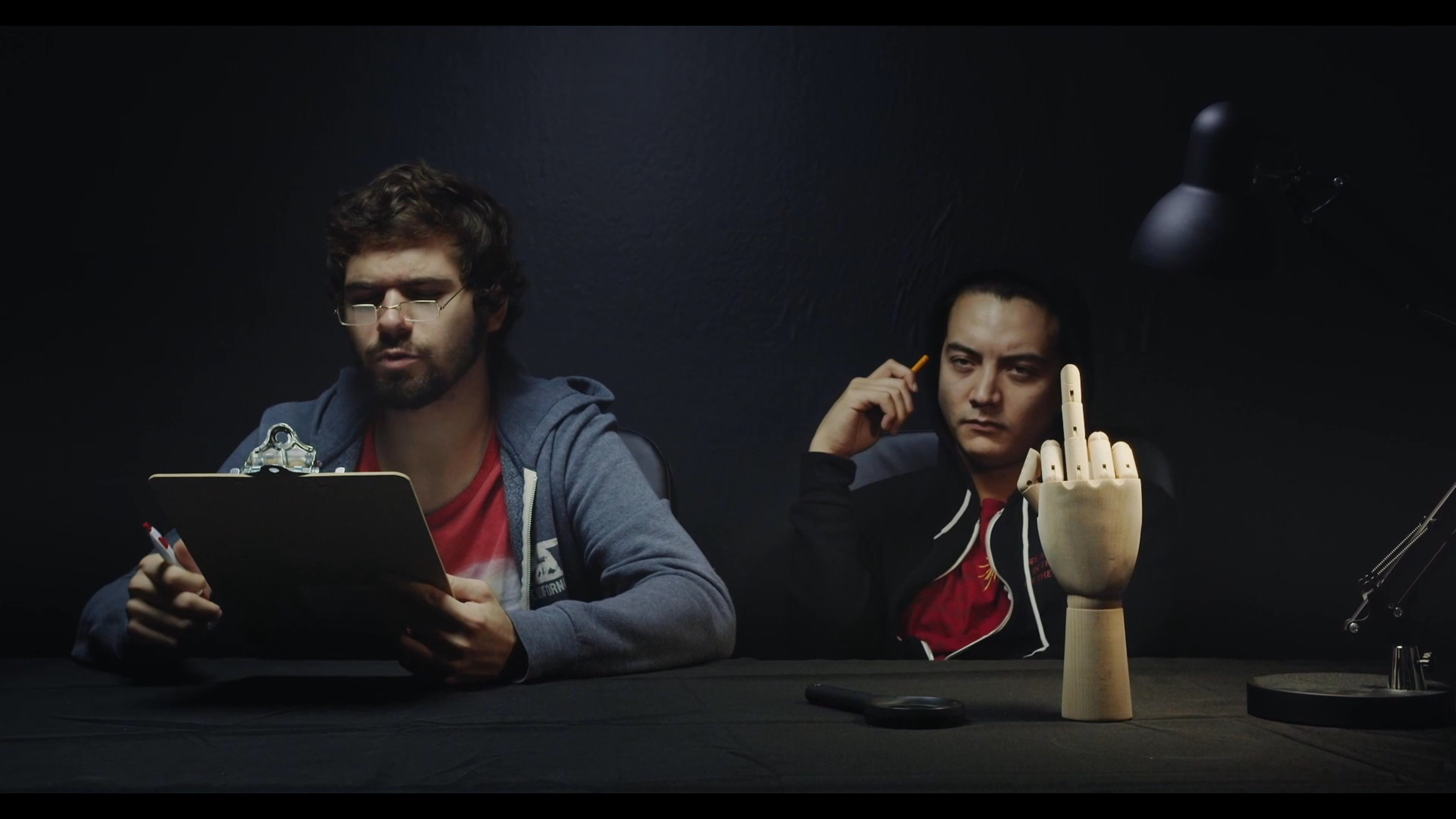 Two individuals engage in a staged conversation within a studio setting, incorporating a wooden mannequin hand as a prop to punctuate their interaction. The scene shifts between an informal discussion and a formal recording environment.