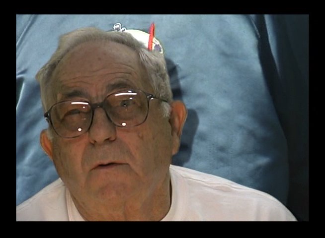 An elderly man named Harry Mayo shares his perspective while participating in an outdoor public gathering. He stands among other attendees in a casual, sunny environment.