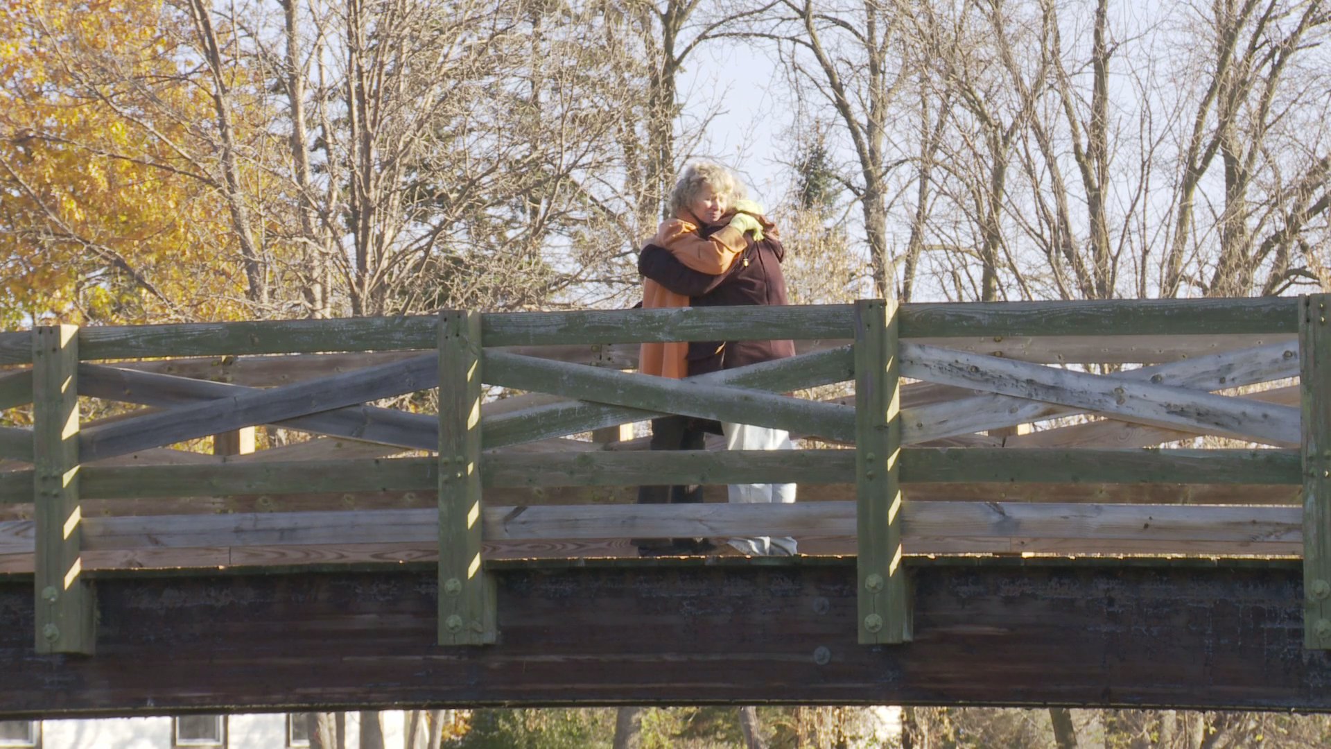 An elderly couple shares an intimate embrace and kiss while standing on a wooden footbridge over a river during a scenic autumn day.