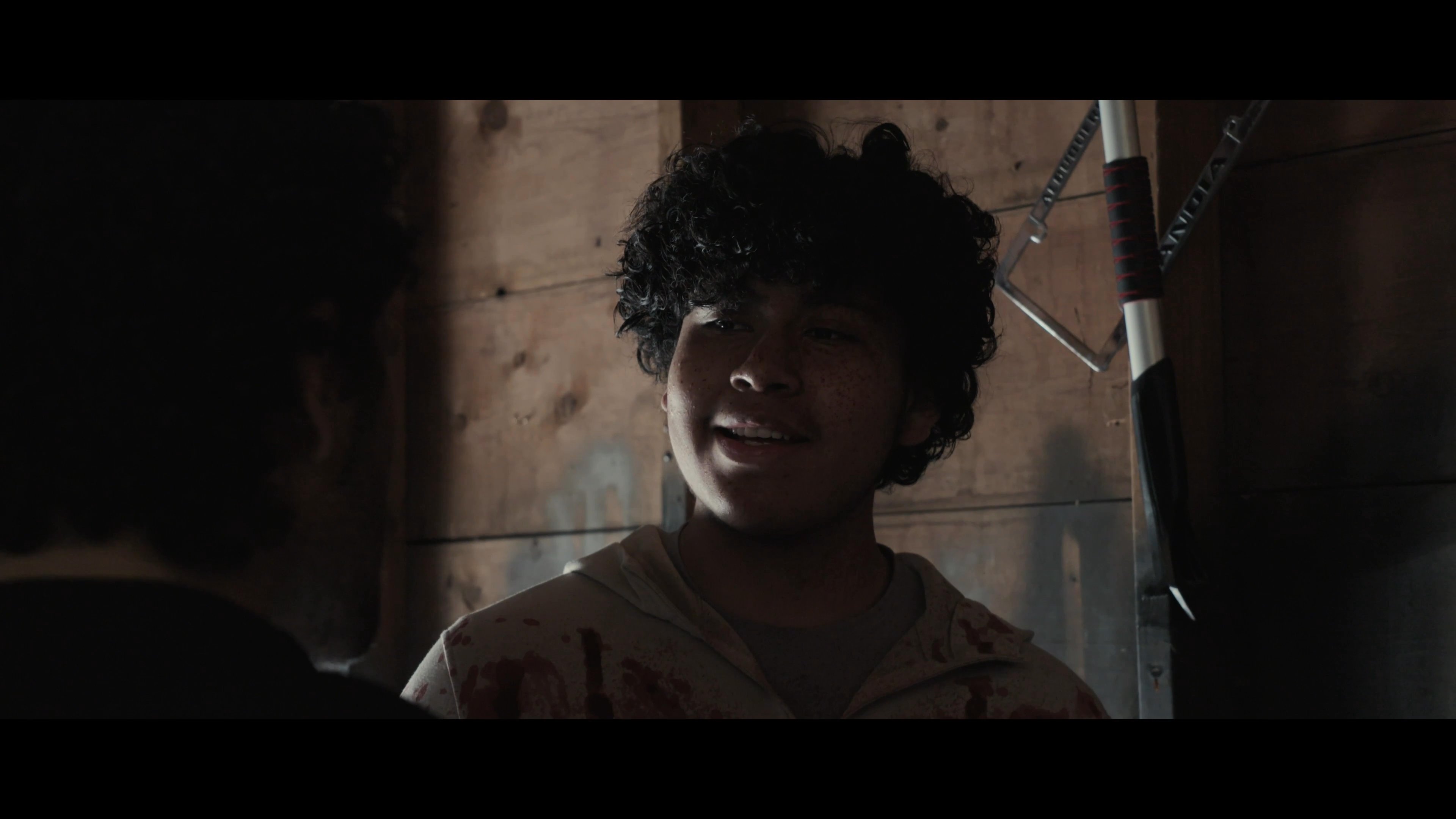 Two men engage in an intense, heated conversation within a dimly lit, wooden-walled space. One man displays signs of physical distress while wearing blood-spattered attire as the pair exchanges urgent words.