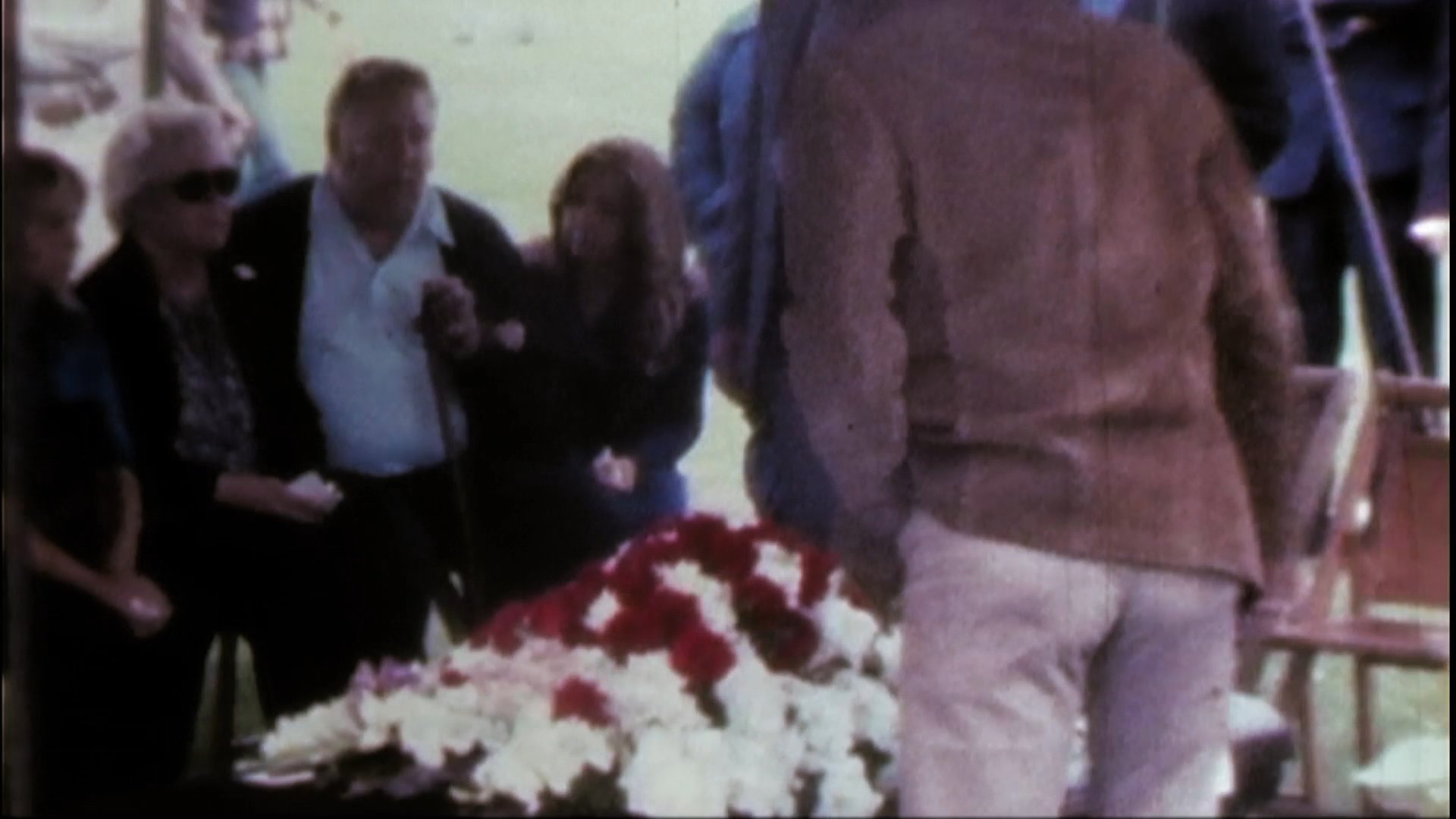 A group of motorcyclists participates in a funeral procession to honor a fallen comrade, culminating in a final tribute at a casket draped in a Harley-Davidson branded flag.