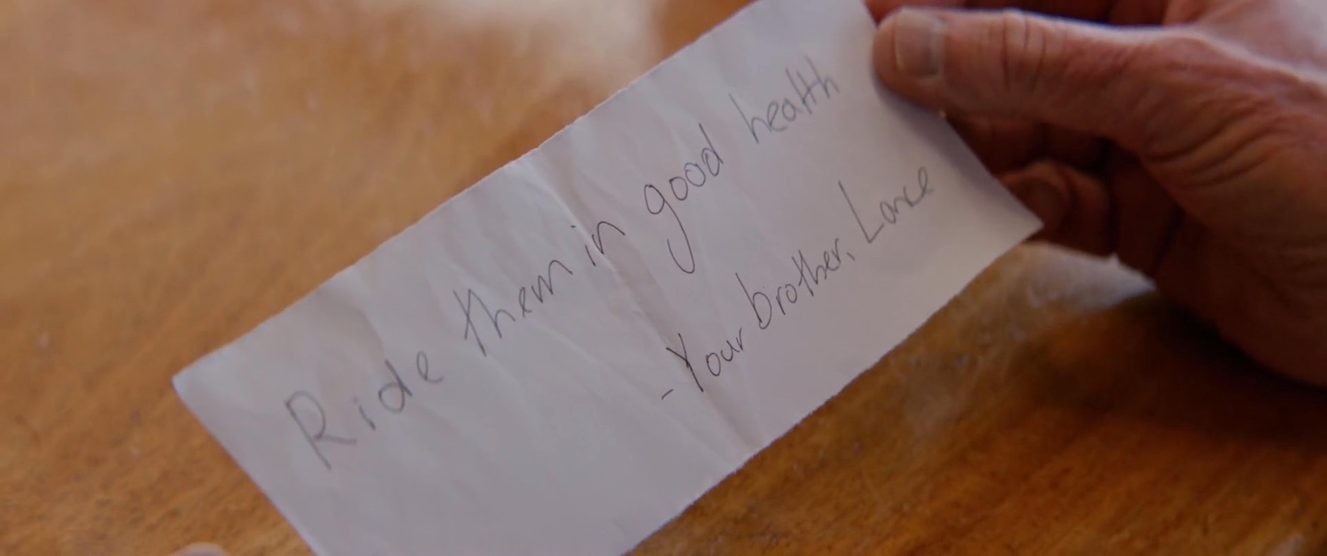 A man receives a surfboard with a handwritten note from his brother, followed by a reflective conversation between two men at a waterfront restaurant.