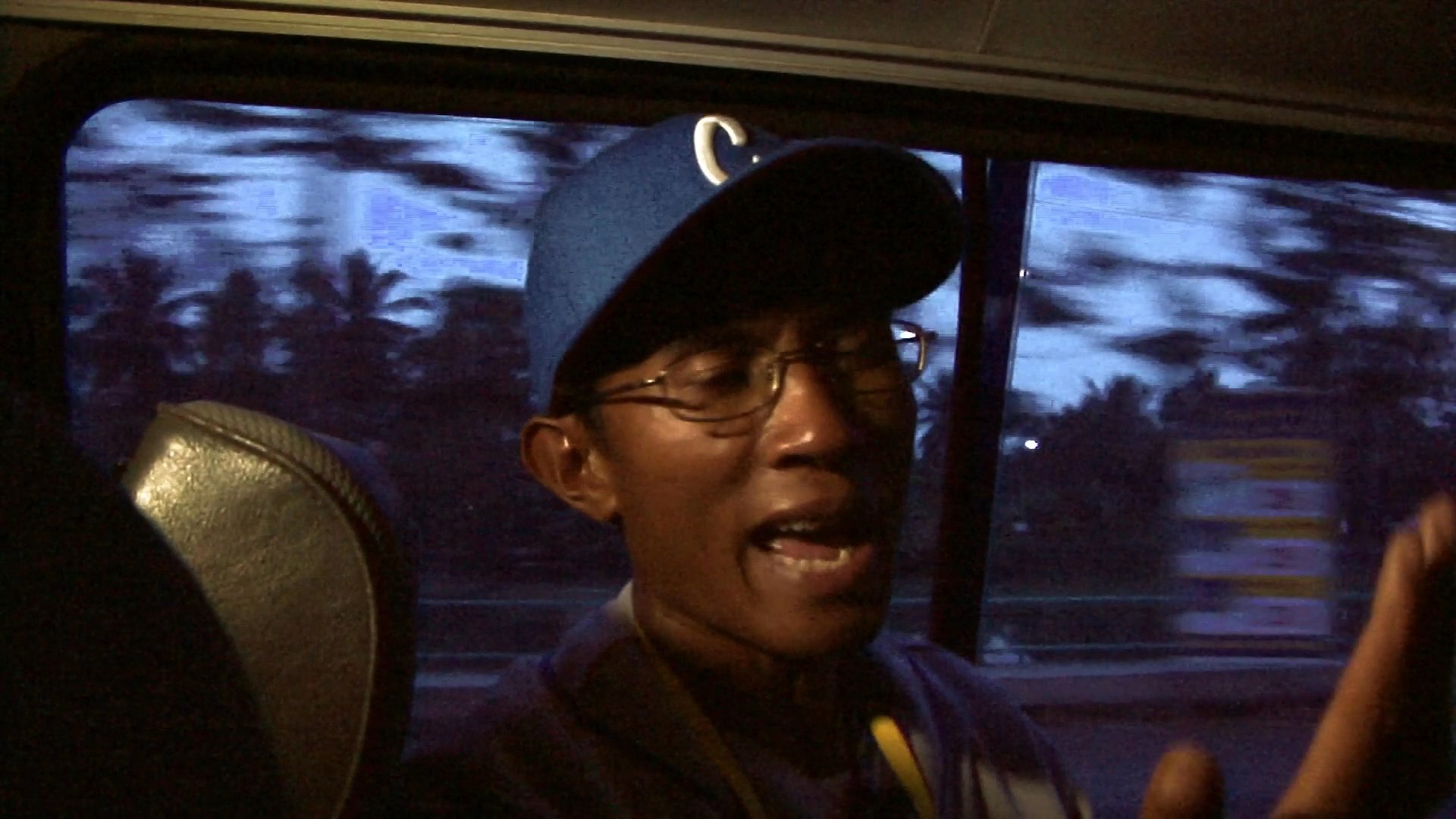 A man wearing a baseball cap speaks while seated in a vehicle traveling along a road at dusk. The blurred landscape outside the window signifies active transit through an urban or suburban environment.