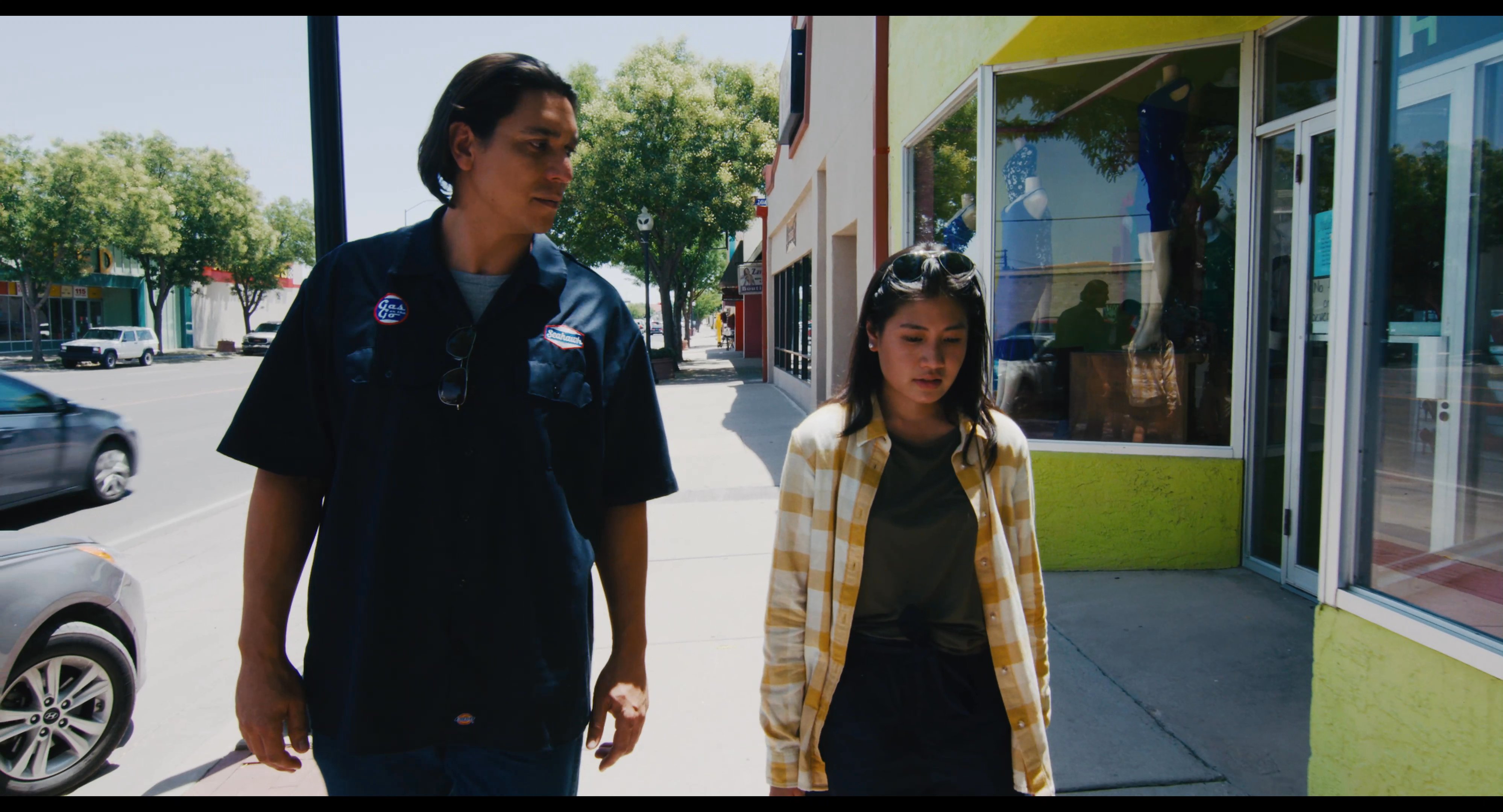 Two individuals walk along a city sidewalk past storefronts while engaging in conversation on a bright, sunny day.