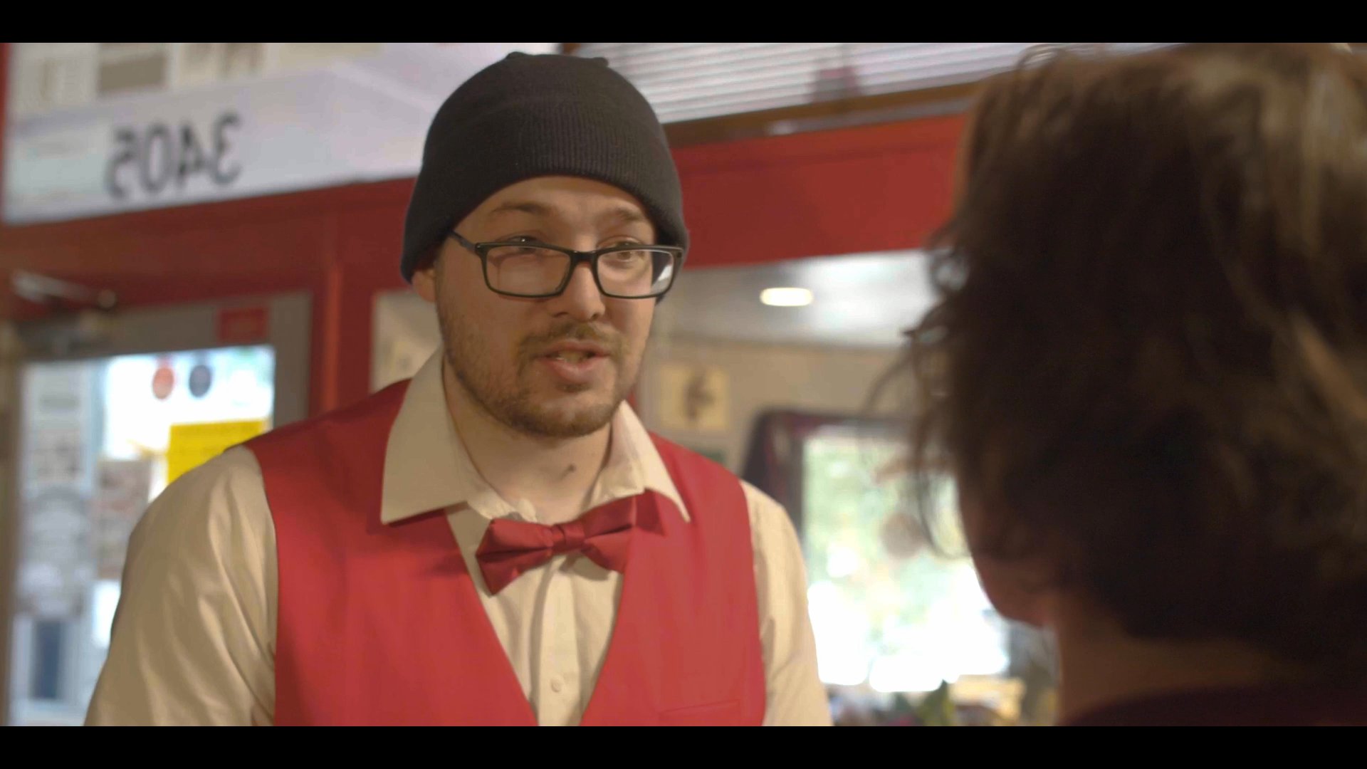 A cinema employee in a uniform discusses a situation with a customer standing at the concession counter. The conversation involves a direct reference to the customer's stomach, prompting an expressive reaction.