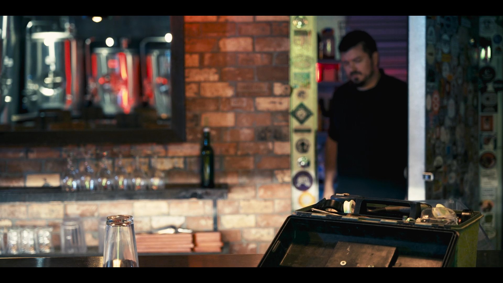 A bartender inspects and adjusts keg equipment in a dimly lit storage area before returning to the bar counter to resume service.