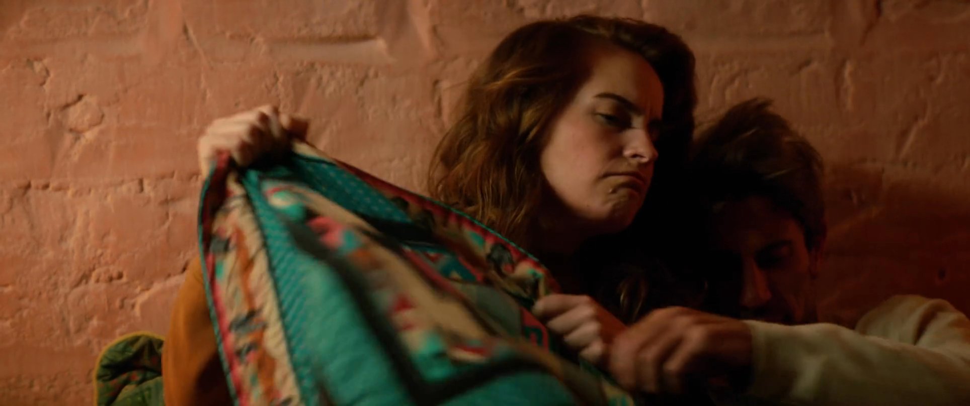 A woman provides physical comfort to a distressed, injured man as they lie together in bed. She pulls the blanket over them and embraces him, offering emotional support in a quiet, intimate moment.
