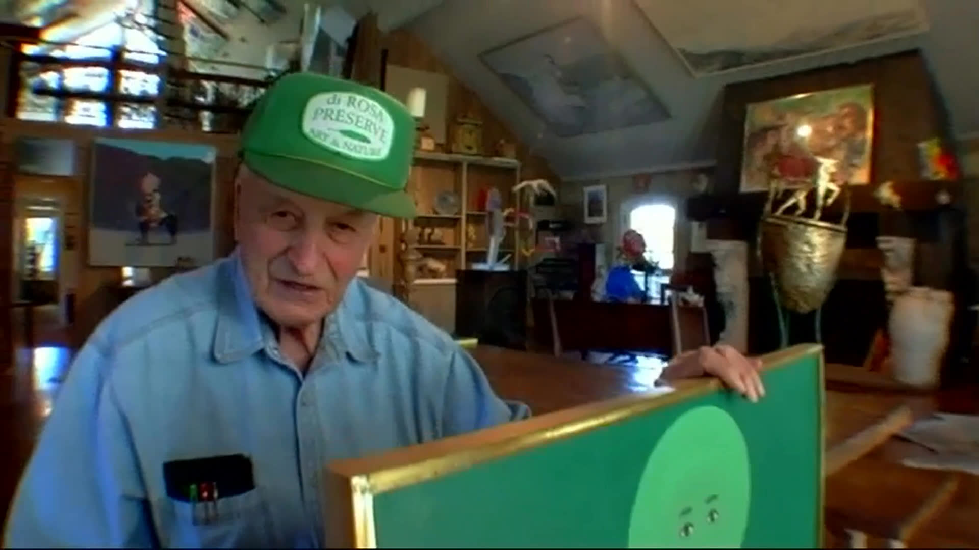 An elderly man in a green cap holds a turquoise painting of a stylized figure in an art-filled room. He recalls being in Europe in 1953, weighing his career and considering writing a great American novel on the Left Bank.