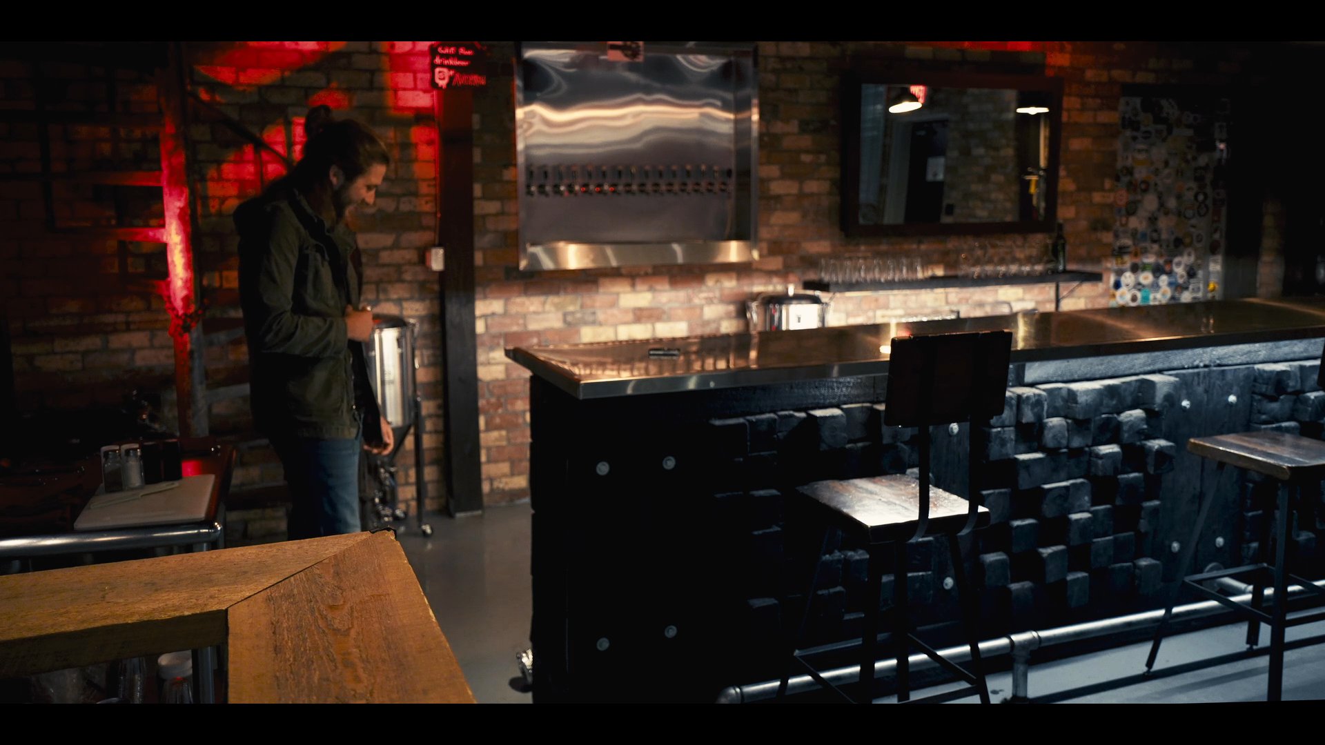 A man moves across a dimly lit, contemporary bar space, eventually settling onto a couch in an adjacent lounge area. The scene utilizes dramatic lighting and a textured brick-and-metal aesthetic to establish a sophisticated, nocturnal mood.