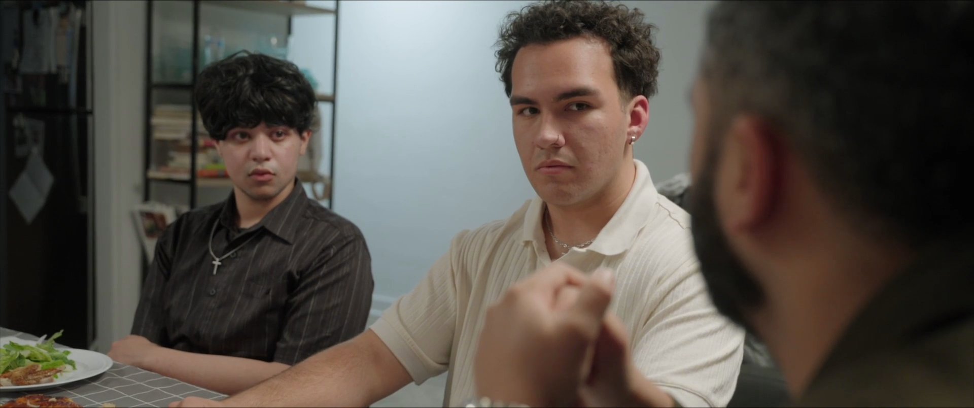 Three men engage in a serious, focused conversation while seated around a dining table. The interaction exhibits a deliberate and contemplative tone as they exchange perspectives during their meal.