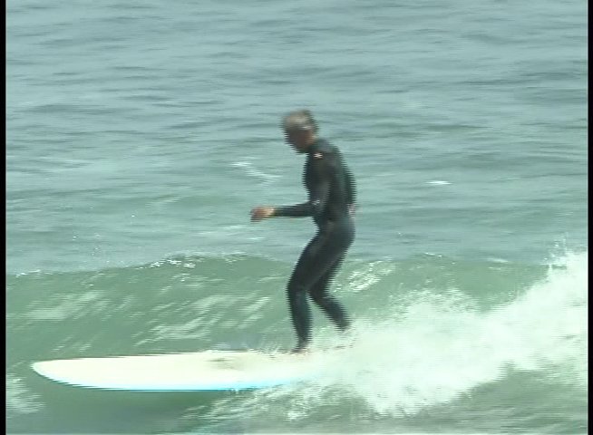 A surfer wearing a wetsuit rides a longboard across a calm ocean wave. The individual maintains balance while gliding smoothly through the water.
