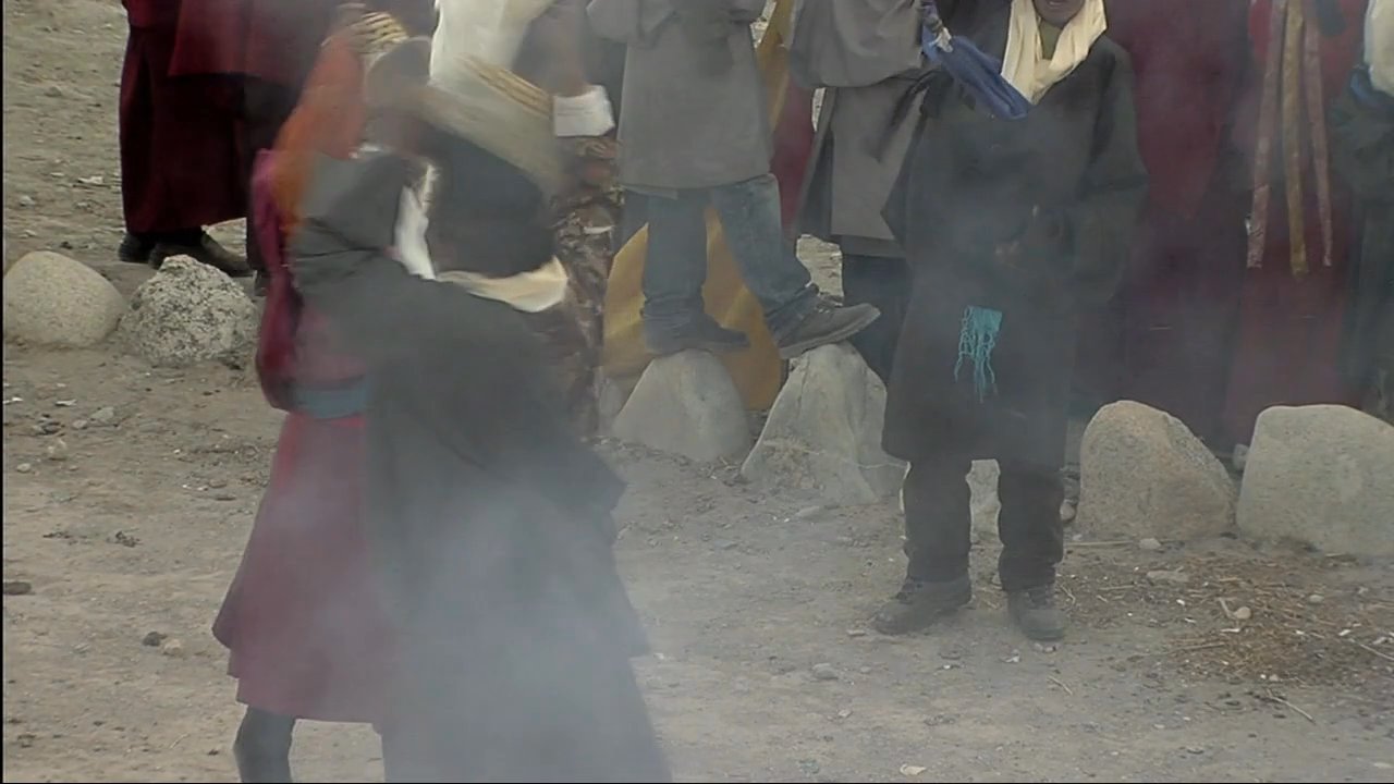 Monks in traditional masks and vibrant robes perform a ceremonial dance in a mountain monastery courtyard while others play long brass horns. The scene highlights cultural traditions set against the dramatic backdrop of the Himalayas.