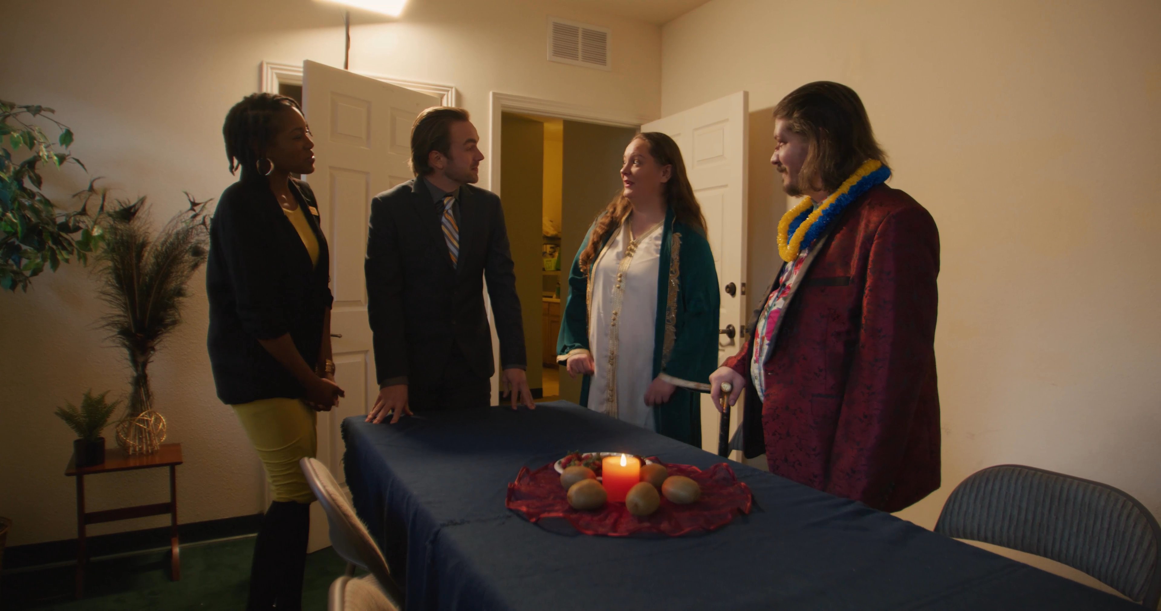 A diverse group of individuals in formal attire convenes in an interior space to socialize and coordinate event details. They exchange greetings and engage in conversation around a table prepared for a small meeting or function.
