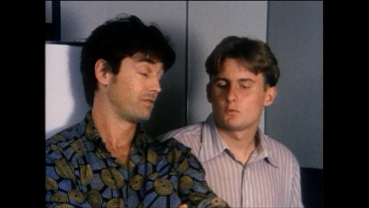 Two men engage in a focused, active conversation within an office environment. The primary speaker gestures while addressing his colleague to explain a point or share information.