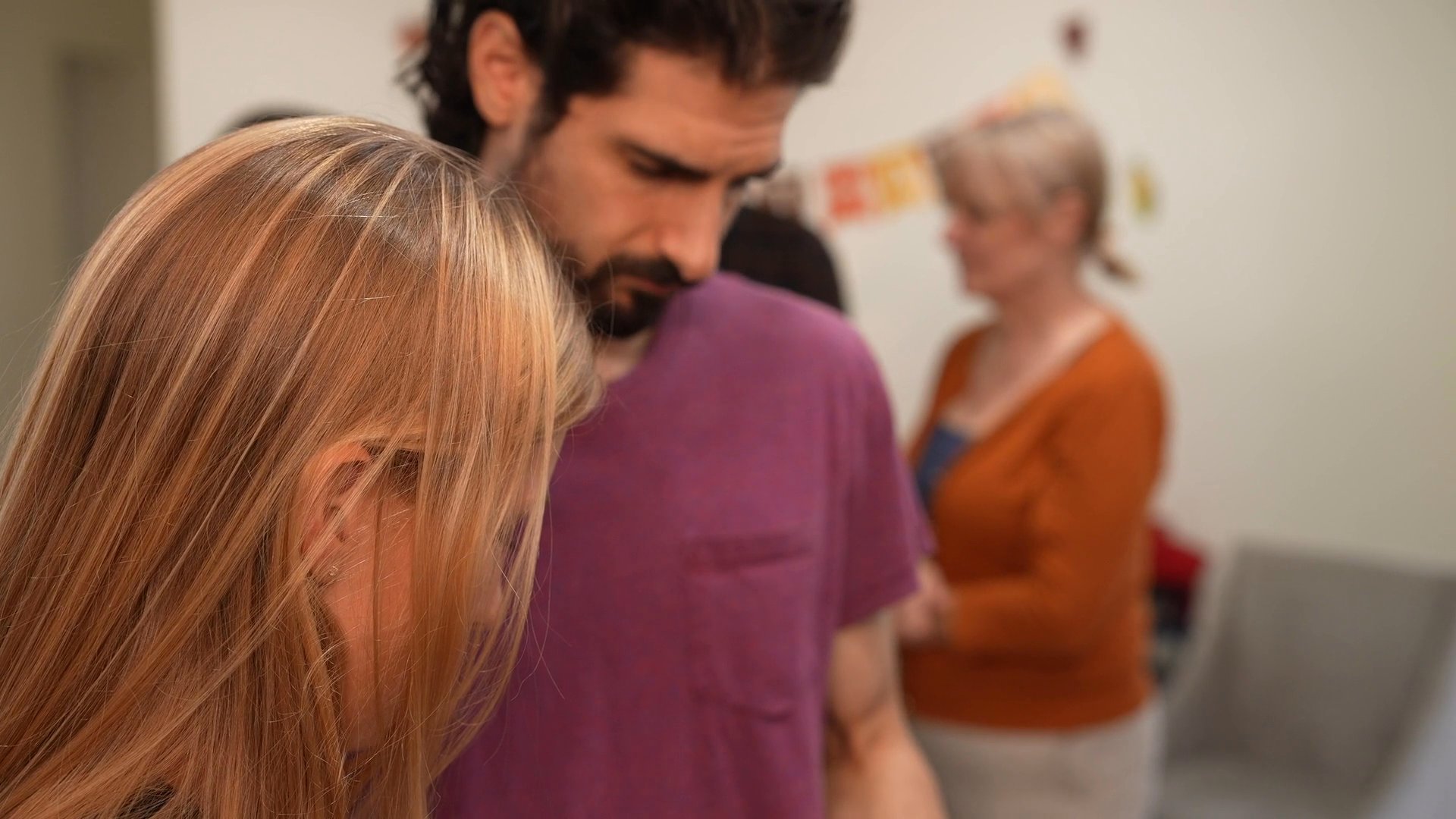 A man and woman engage in a focused conversation while standing in a room with other guests in the background.