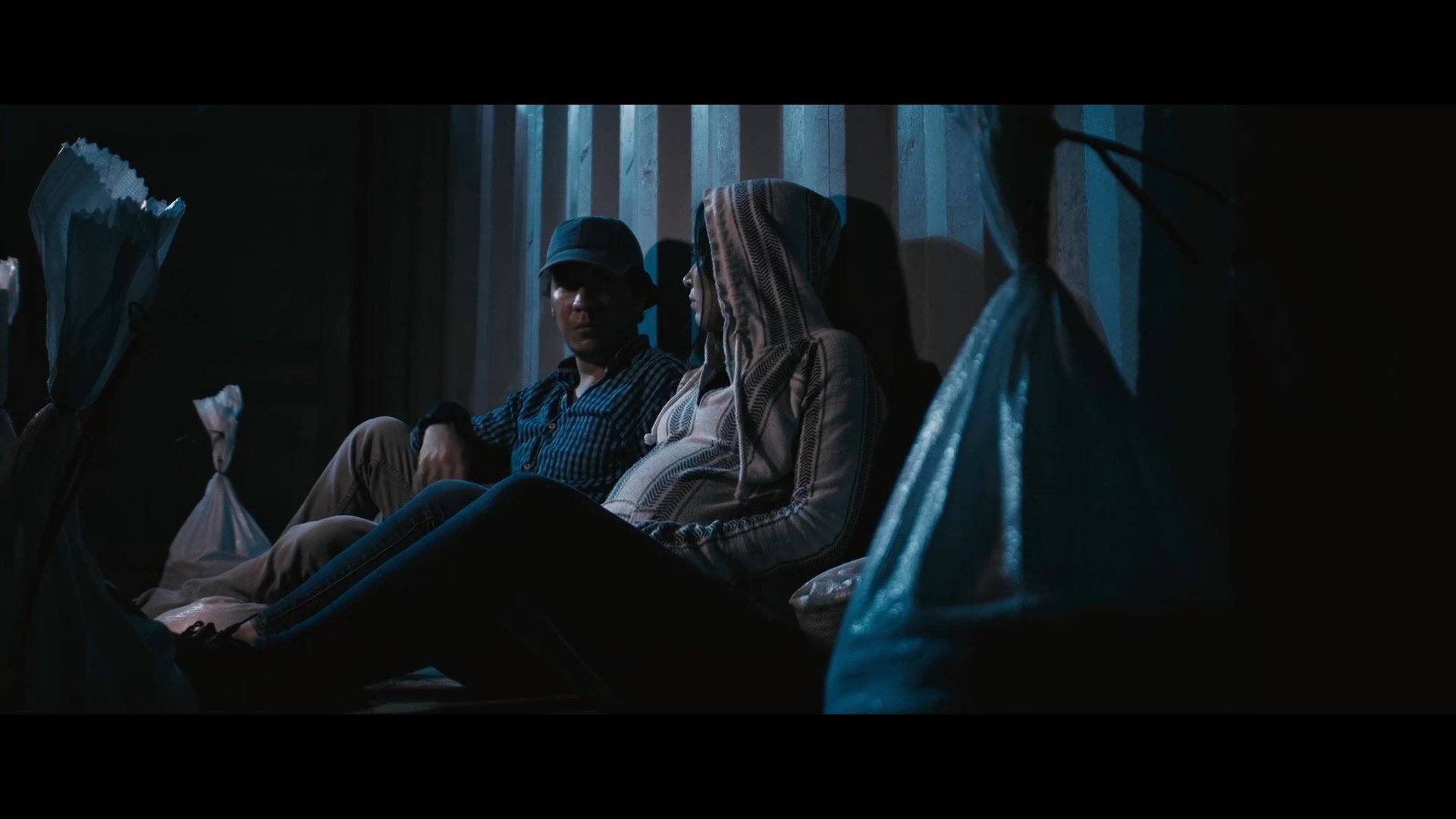 A man and a pregnant woman sit in a dimly lit, austere room while discussing the stress of their financial instability. The conversation highlights their shared anxiety regarding their lack of funds and uncertain future.