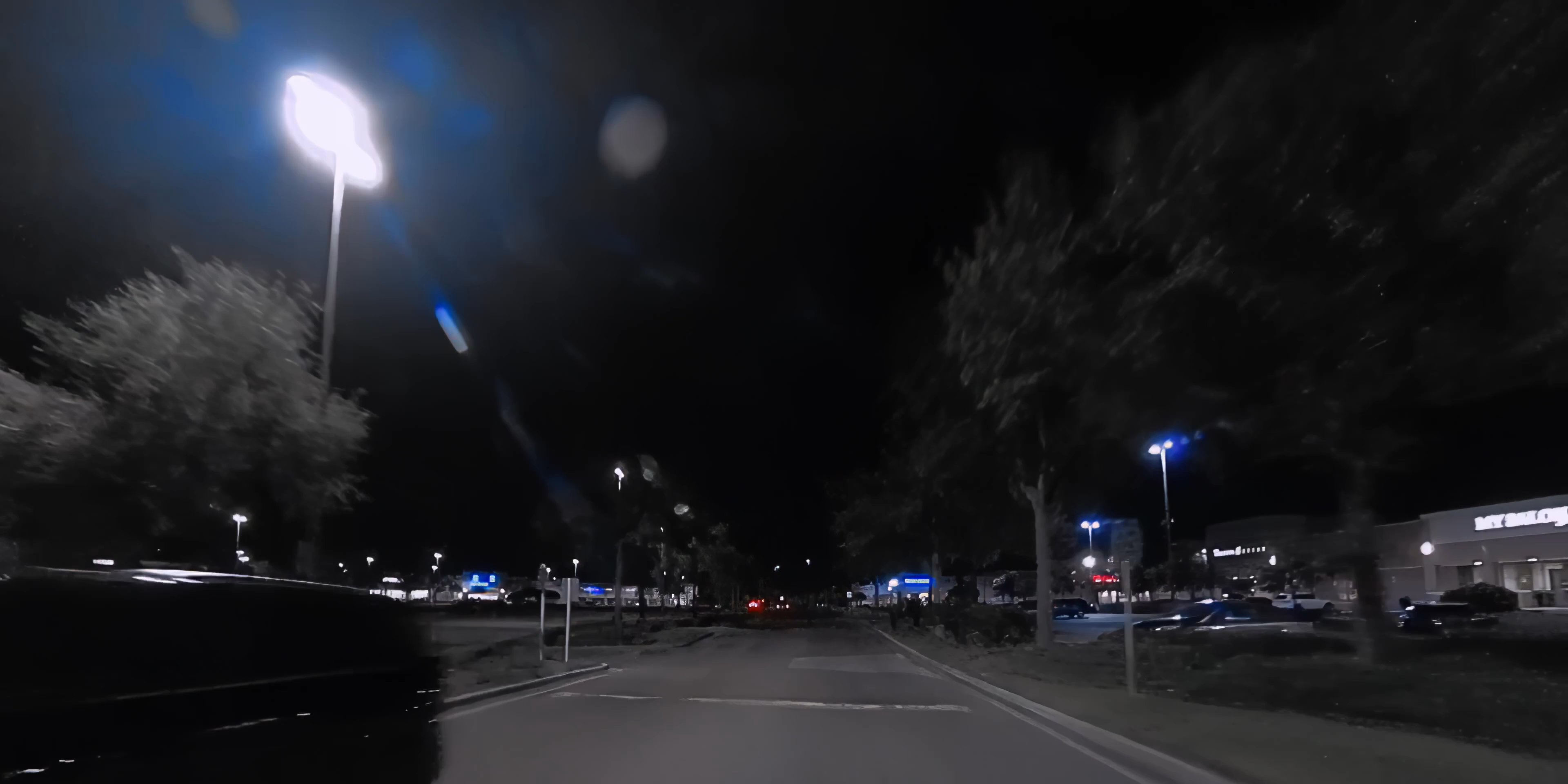 An aerial perspective captures a coastal city at twilight before transitioning to a ground-level view of a vehicle moving through a brightly lit commercial parking area at night.