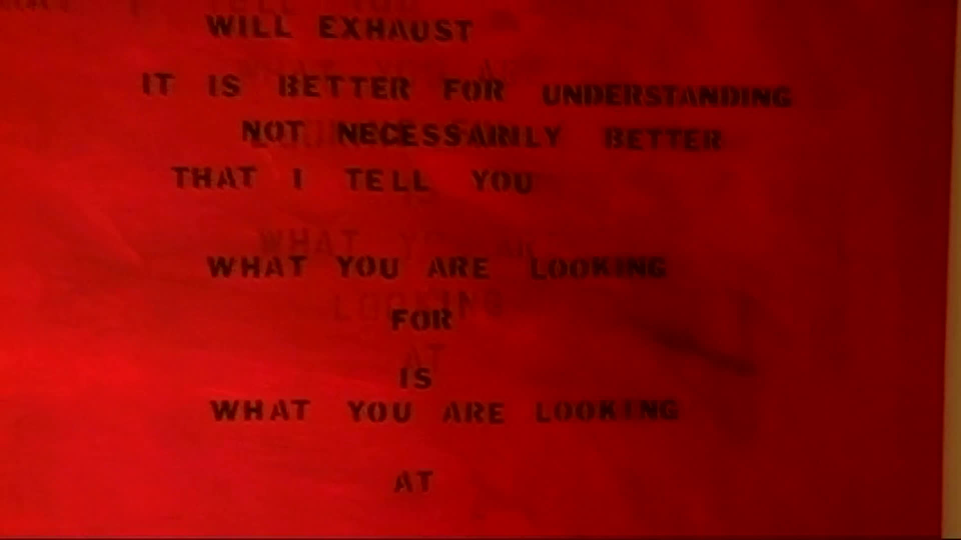 A red text-based artwork fills the gallery, with close views of its layered, poetic wording. An older man and a young woman speak beside the installation in a warmly lit exhibition space.