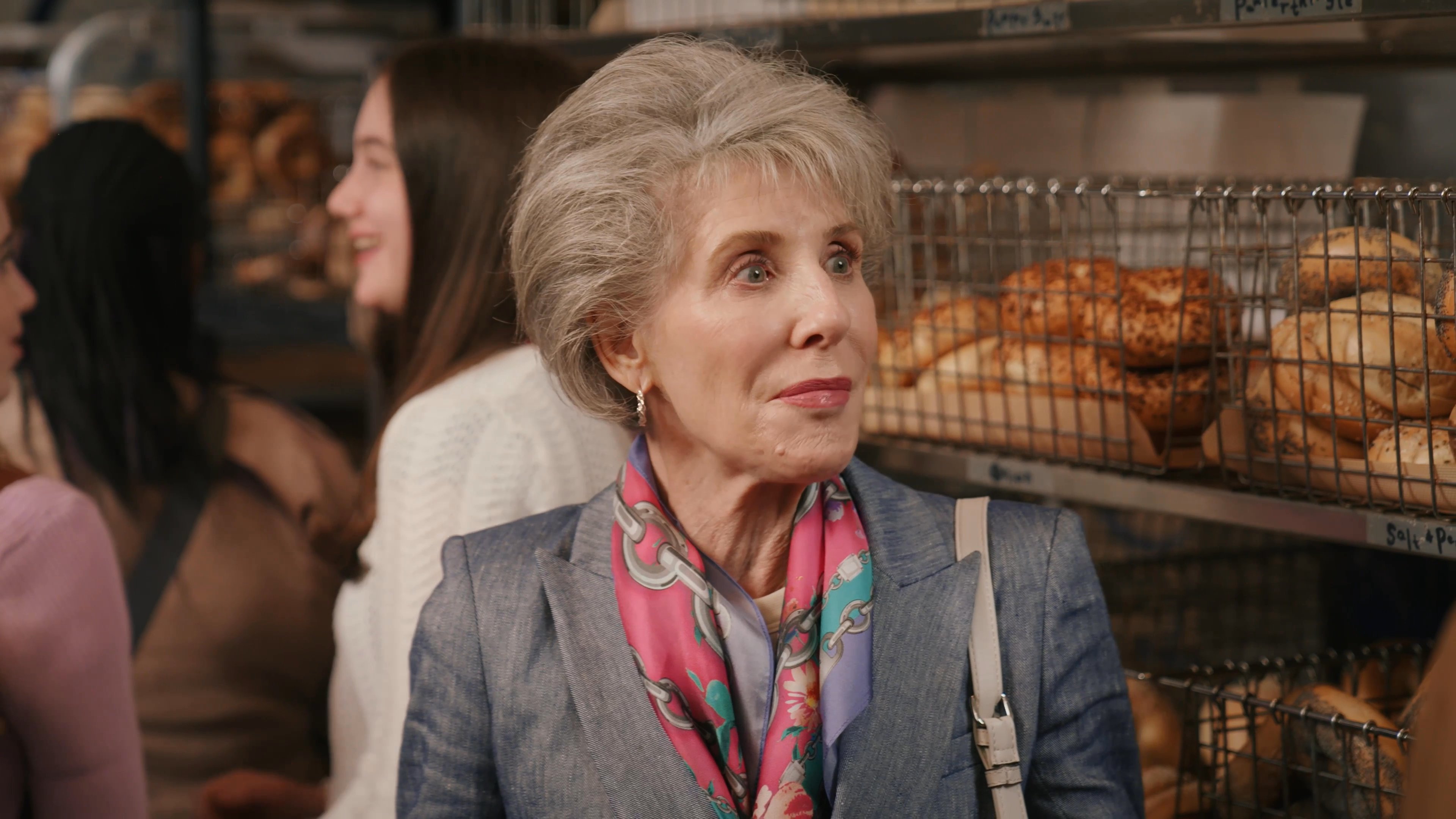 An older woman and a young man engage in a friendly conversation while standing in a busy bakery shop. The interaction occurs amidst other customers waiting in line.