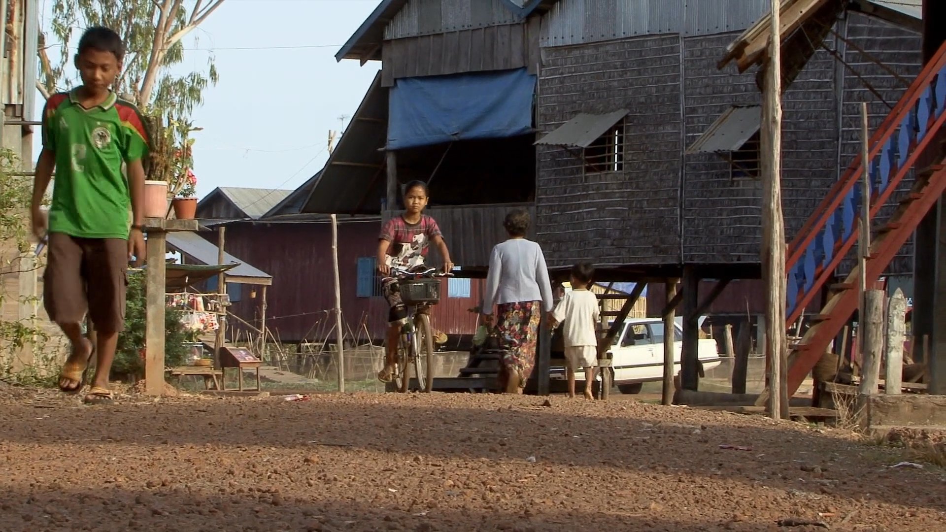 Local villagers engage in agricultural tasks and transportation in a rural setting, featuring individuals watering garden plots and a parent cycling with three children.