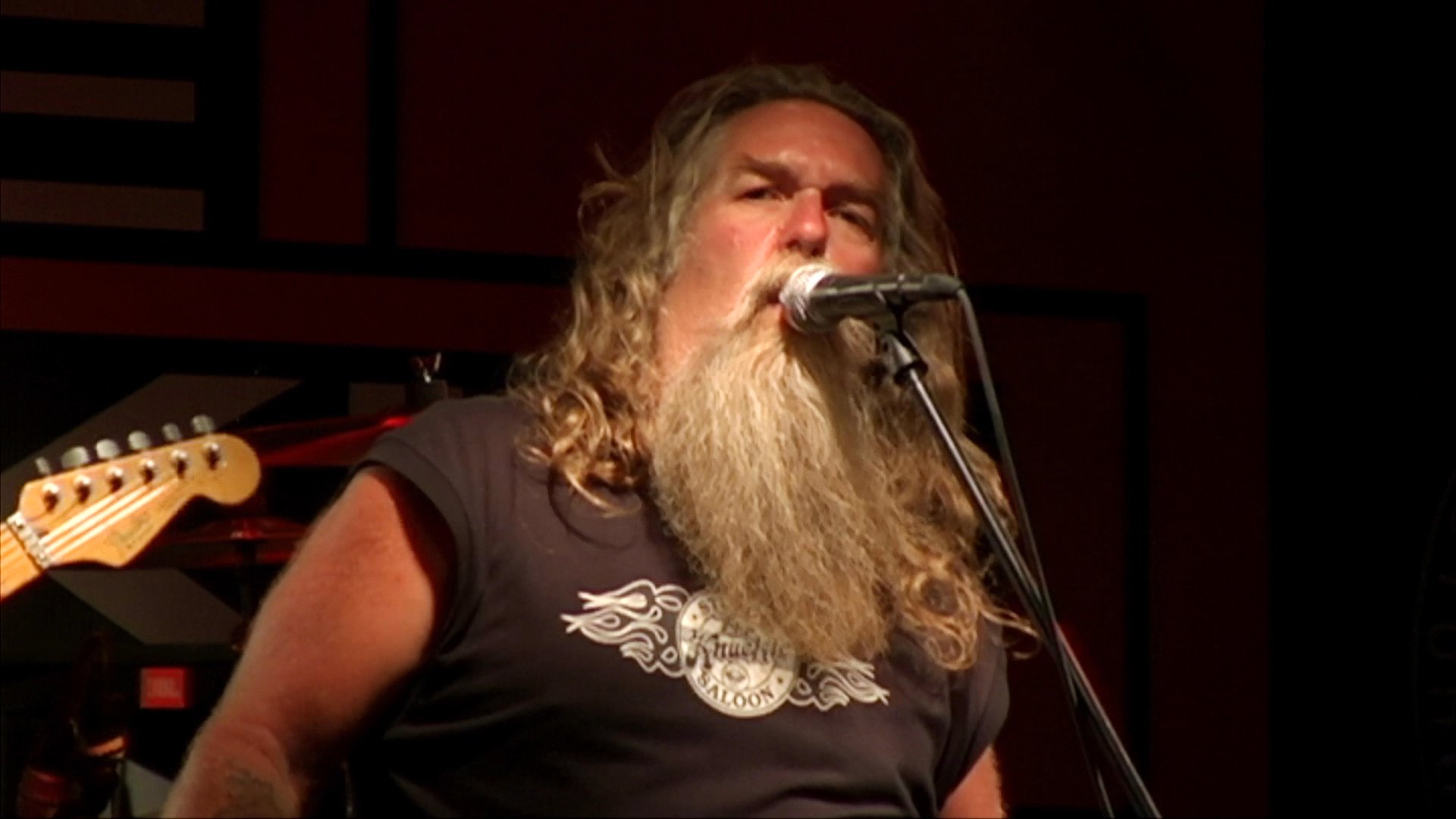 A long-haired vocalist performs on stage at a crowded bar while patrons dance and socialize on the floor.
