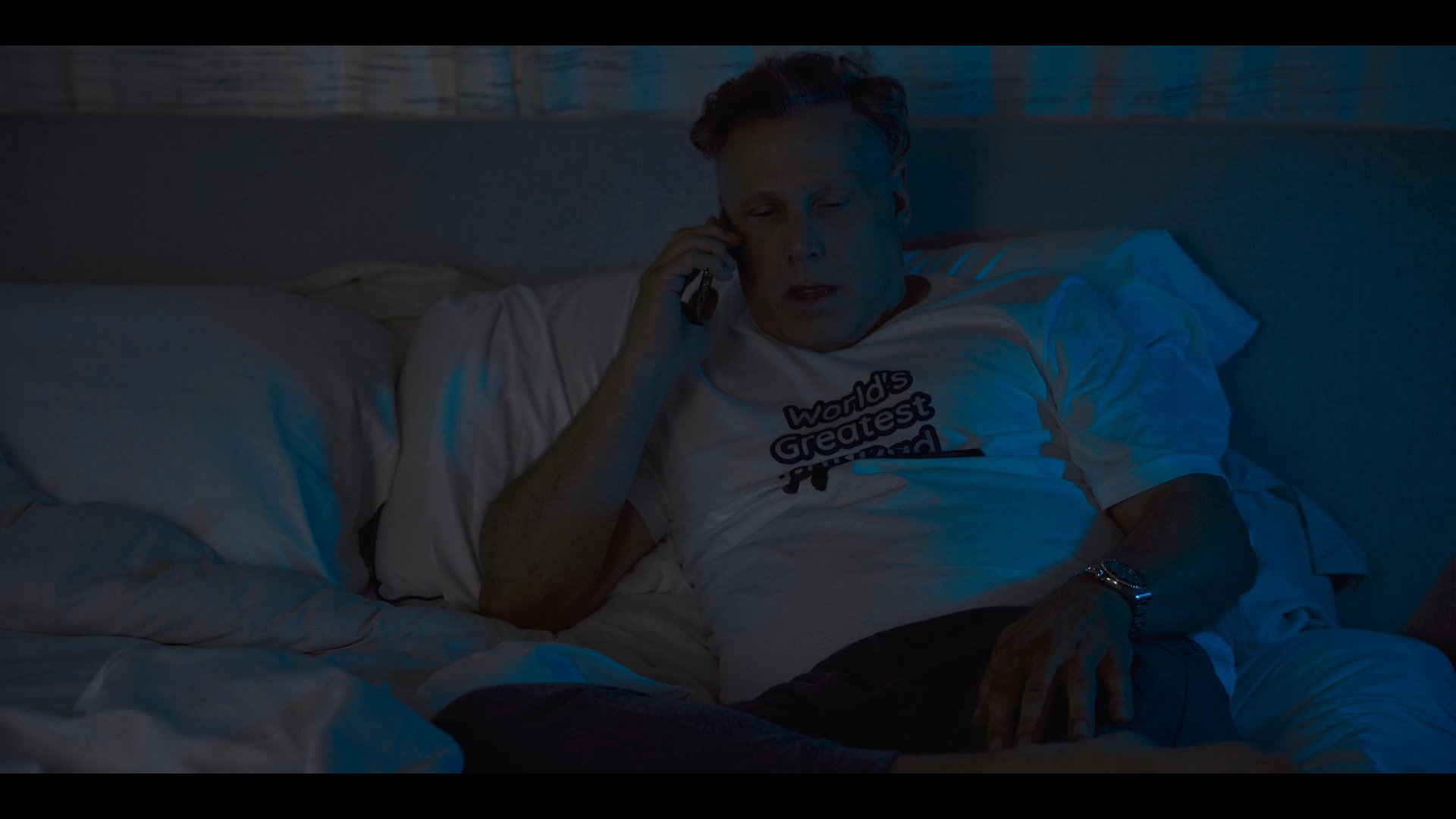 A man sits in bed at night having a conversation on his mobile phone. The dimly lit room suggests a late-night setting.