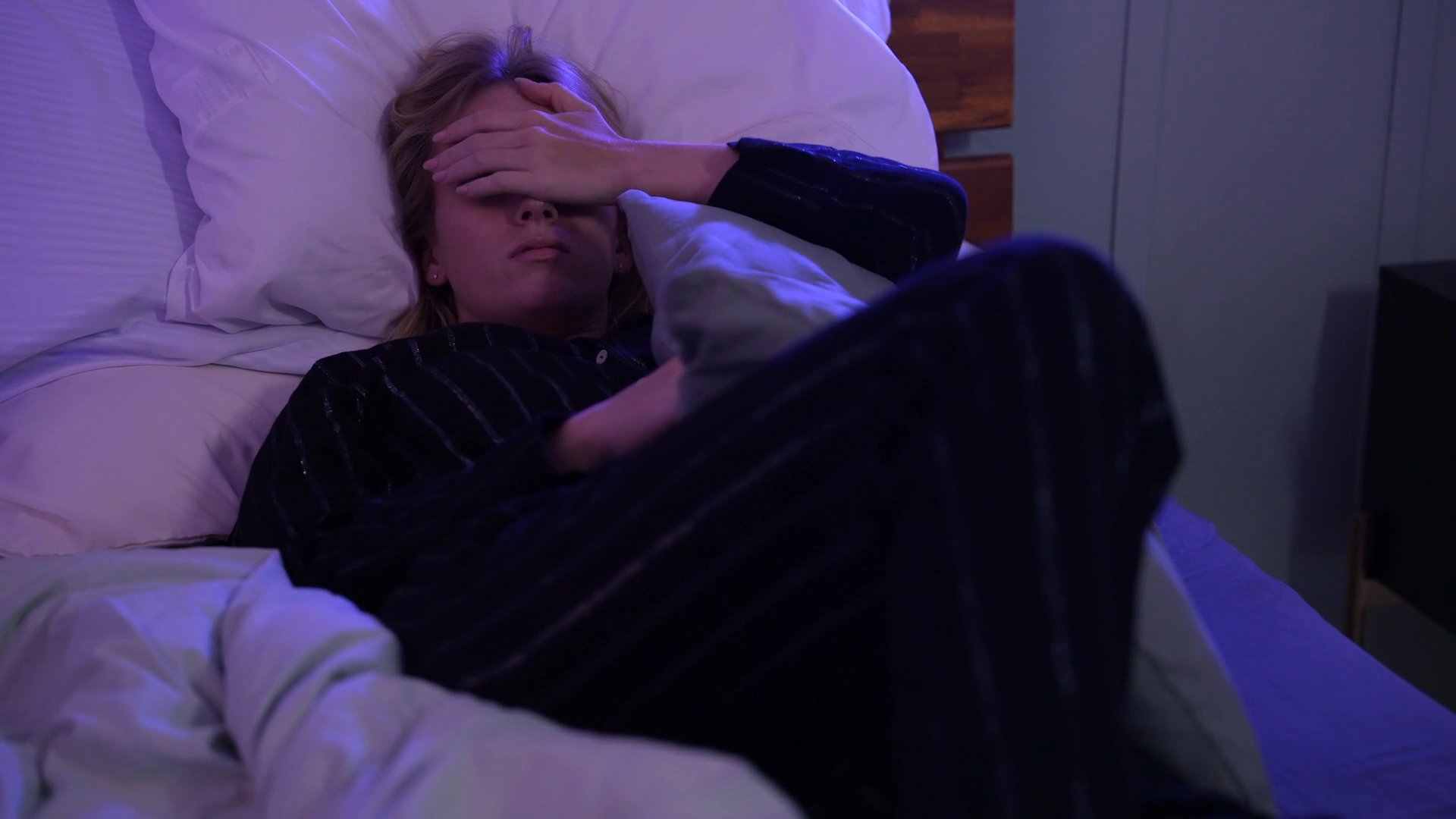 A woman struggles with sleep in her bedroom during the night, showing signs of distress as she wakes and presses her hand to her forehead. She eventually observes a partner sleeping beside her while remaining awake and unsettled.
