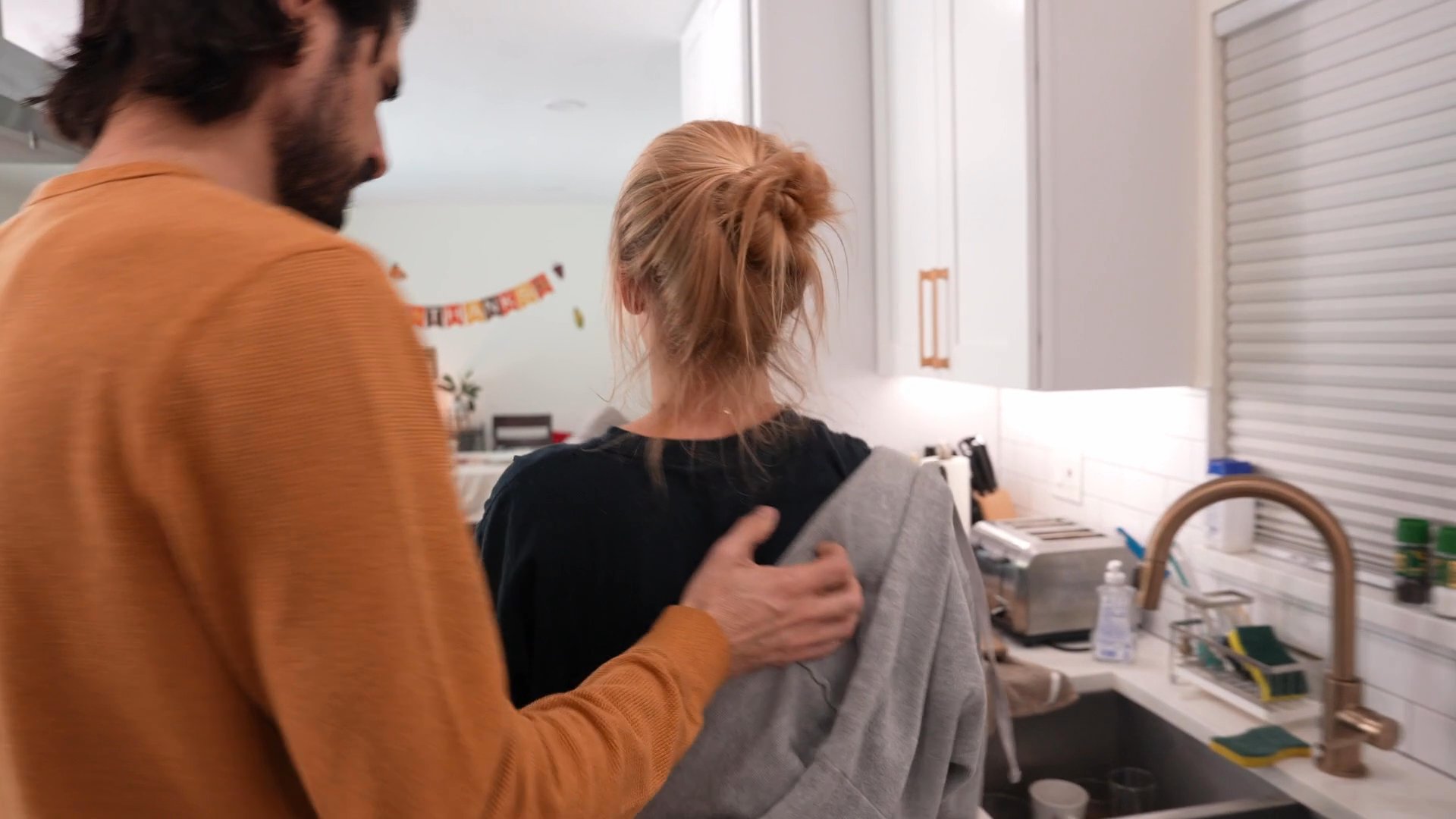 A woman experiences social discomfort while navigating a gathering in her home as guests interact around her. The scene captures the shift from chaotic group dynamics to her quiet withdrawal from the living area.
