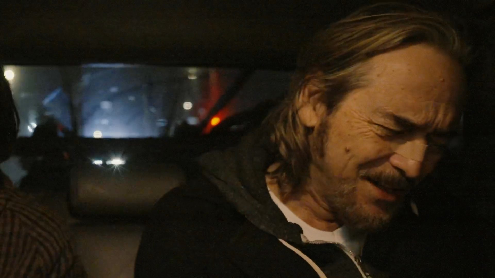 Two men and a woman engage in a tense conversation while seated inside a parked car at night. The dialogue shifts between concern and erratic behavior as the group navigates an uneasy situation.