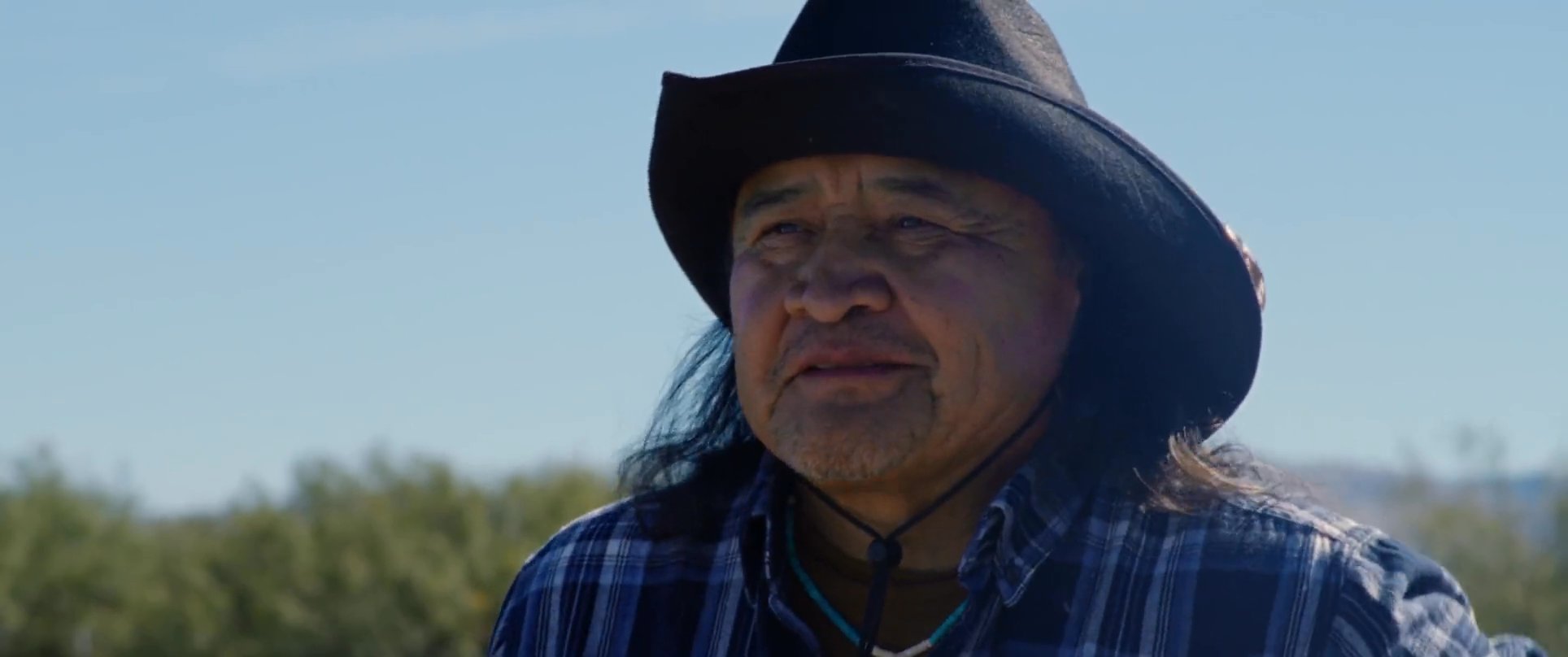 A man in a white t-shirt engages in a serious discussion with an experienced rancher amidst an open agricultural field. The two exchange perspectives regarding land management or operational planning under clear daylight conditions.