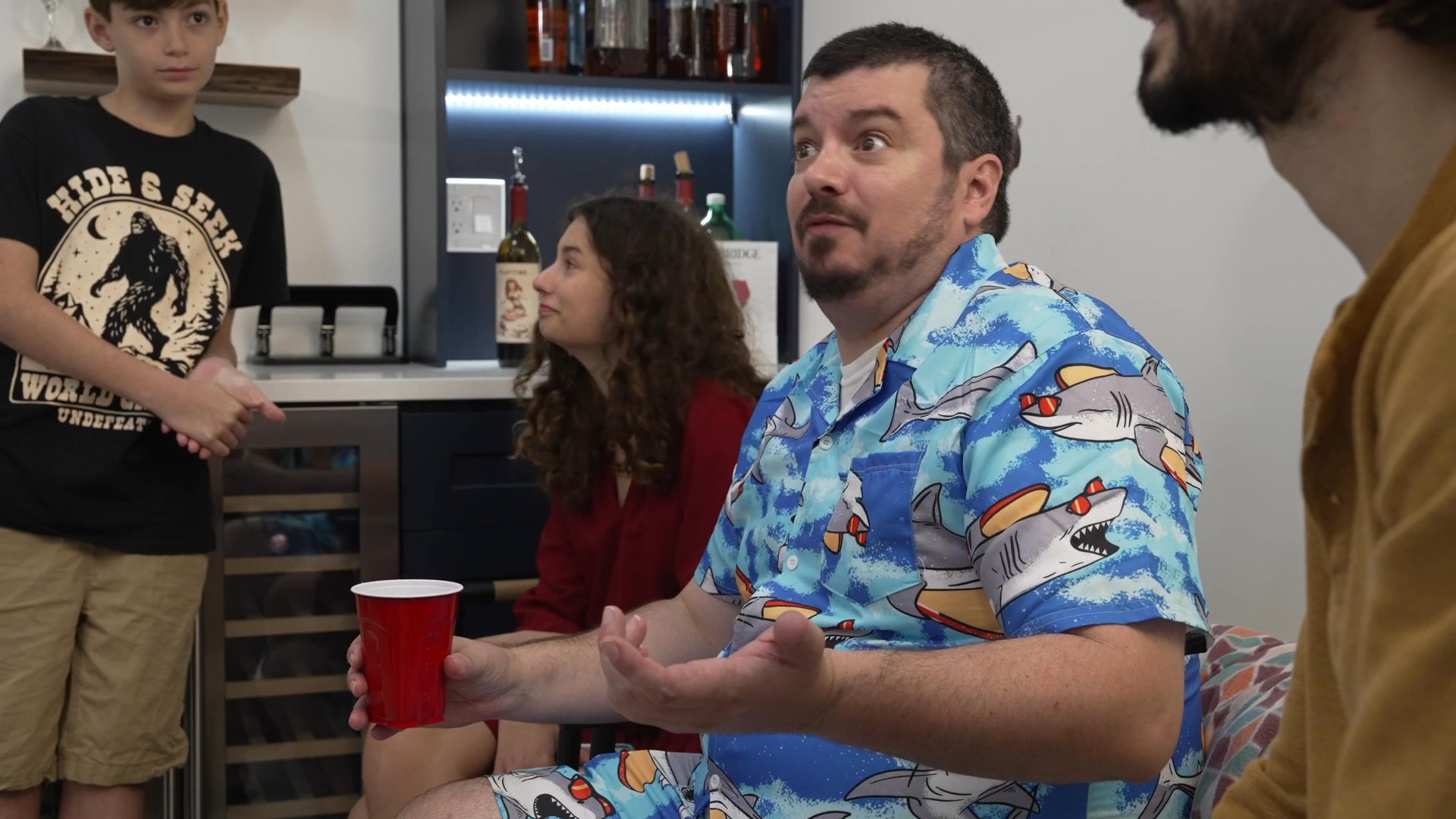 Participants engage in an animated group discussion within a residential home environment. The individuals utilize expressive hand gestures and dialogue to share perspectives in a casual social setting.