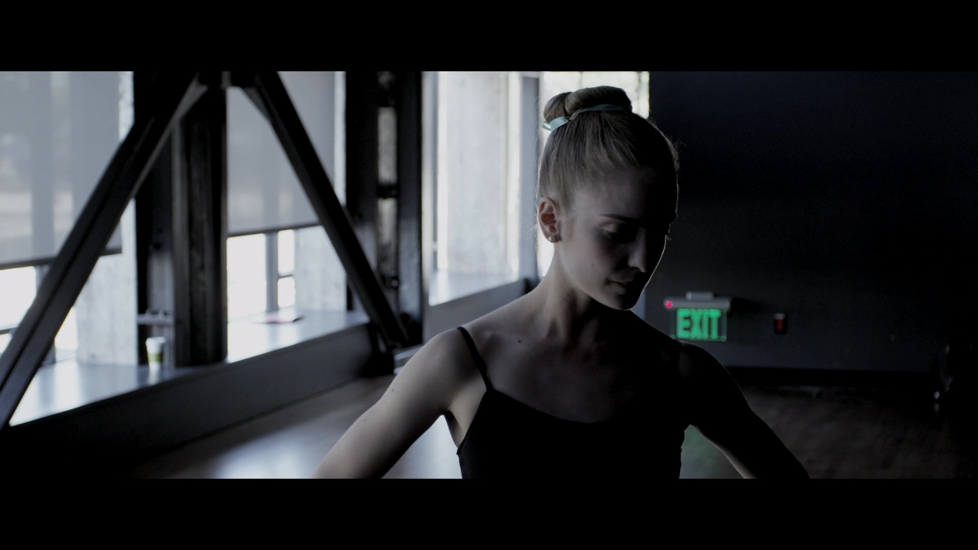A focused ballerina performs a choreographed routine under the guidance of an instructor in a professional dance studio. The session emphasizes precision and movement as the dancer executes steps in silhouette against expansive window lighting.