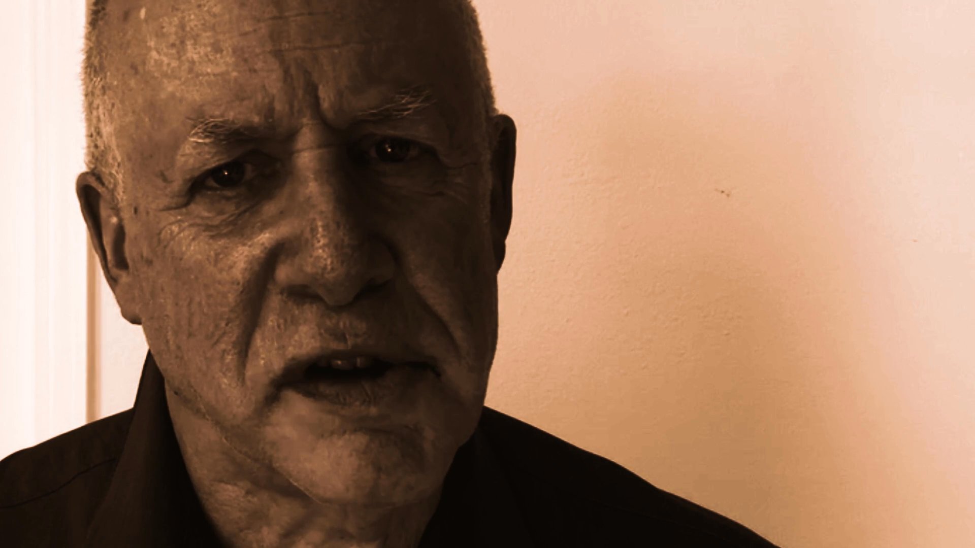An older man delivers a spoken address, maintaining a serious and focused demeanor throughout the sequence. The monochromatic sepia filter emphasizes a contemplative and direct presentation.