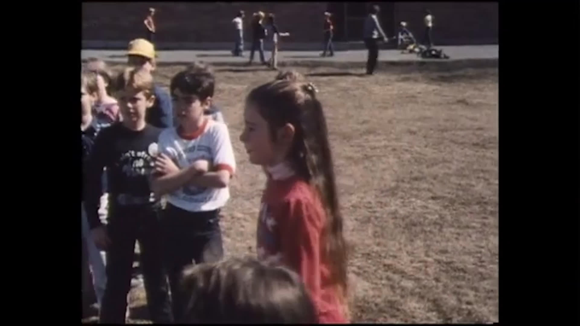 Two boys talk about town girls being prettier and having fancier things while a group of children gathers on a dry outdoor field. A girl in red tosses a yellow ball as the others watch and play nearby.