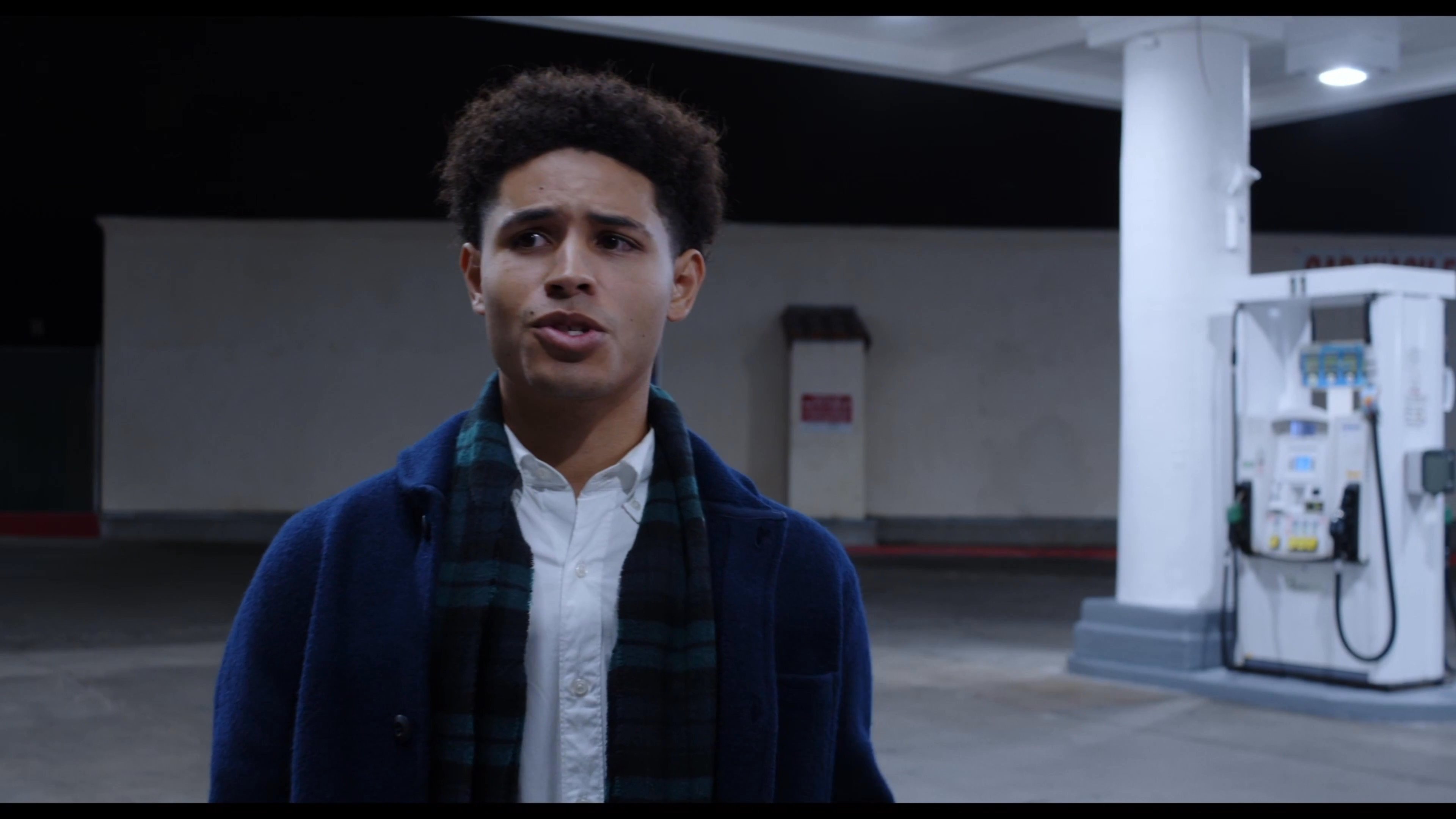 Two young men engage in a relaxed conversation while standing at an outdoor gas station at night. One individual snacks on a food item while the other listens attentively in the well-lit, quiet environment.