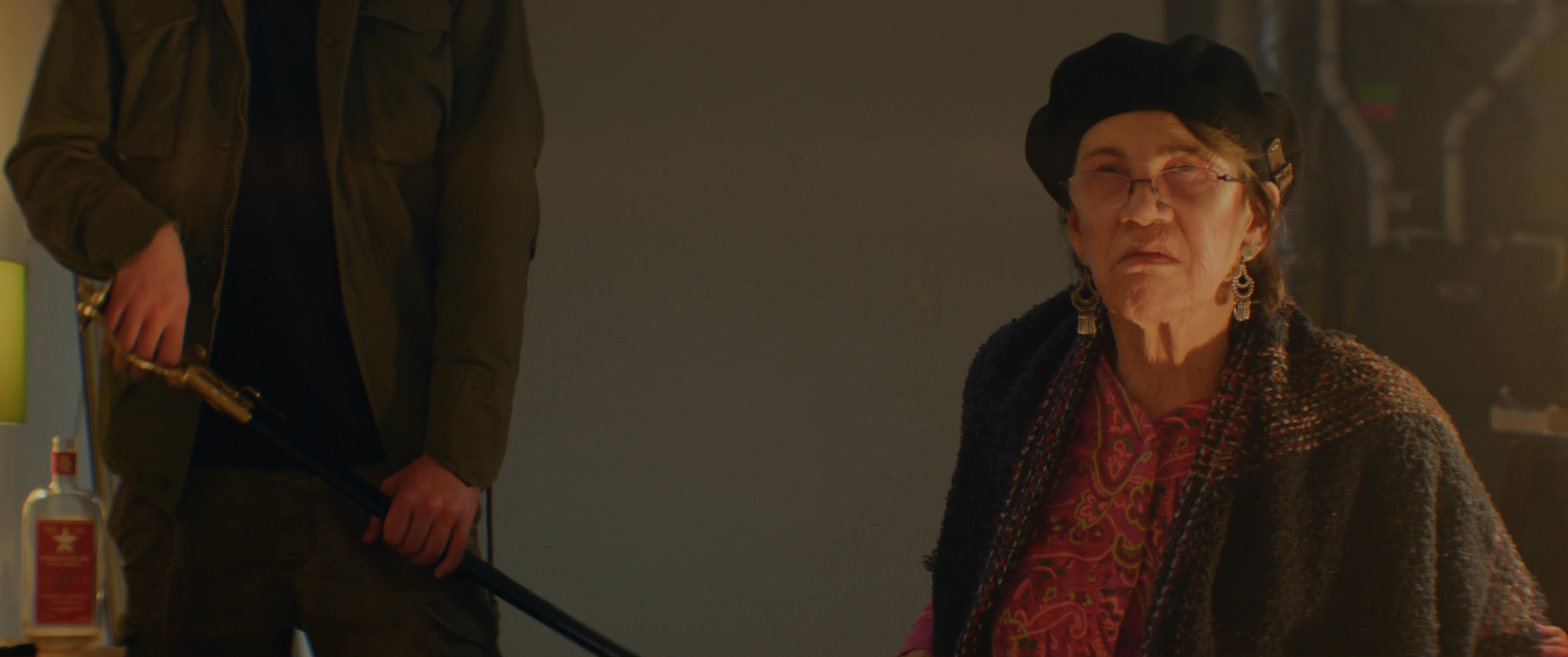 An elderly woman wearing a beret engages in a tense dialogue with a young man in a room decorated with protest posters. The interaction appears to be part of a rehearsed dramatic scene.