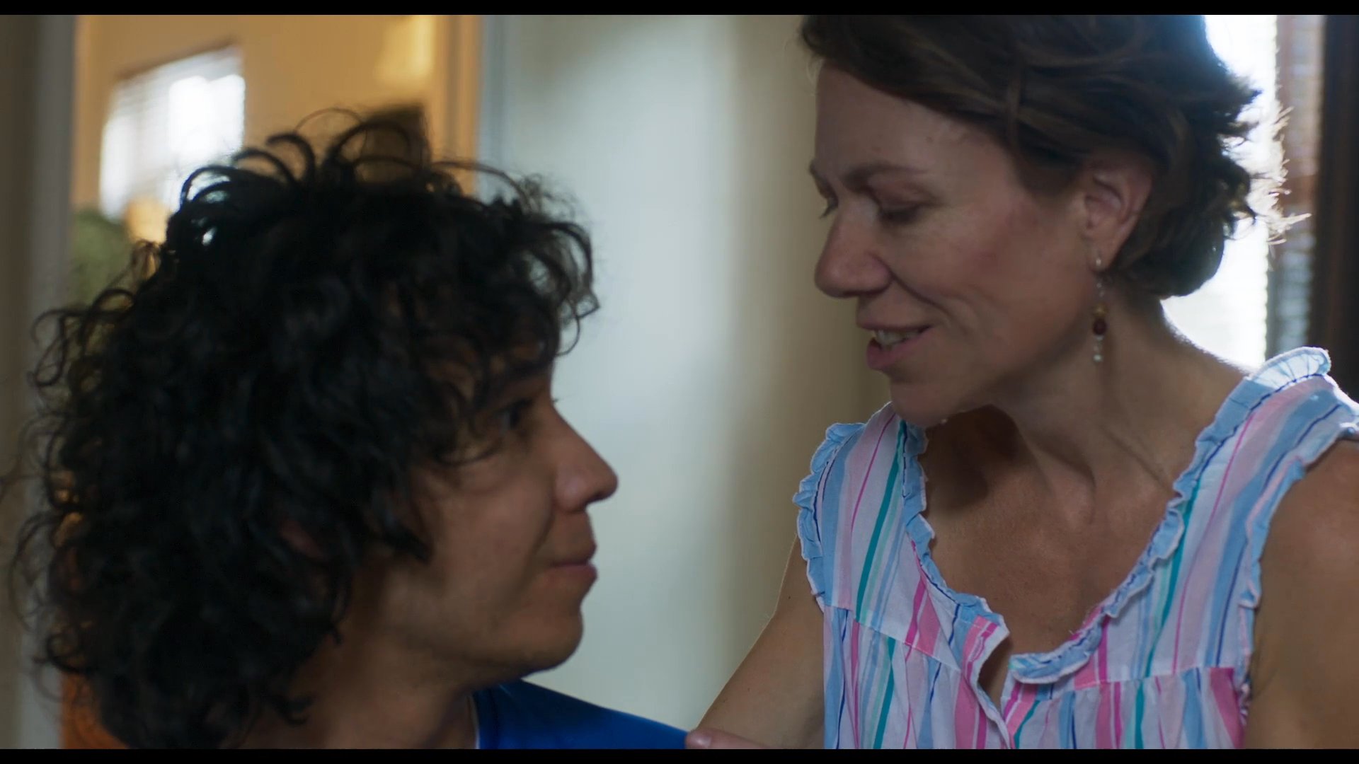 A woman leans toward a young man sitting on a bed to share a tender, affectionate moment in a bedroom. The scene emphasizes their close emotional connection and personal interaction.