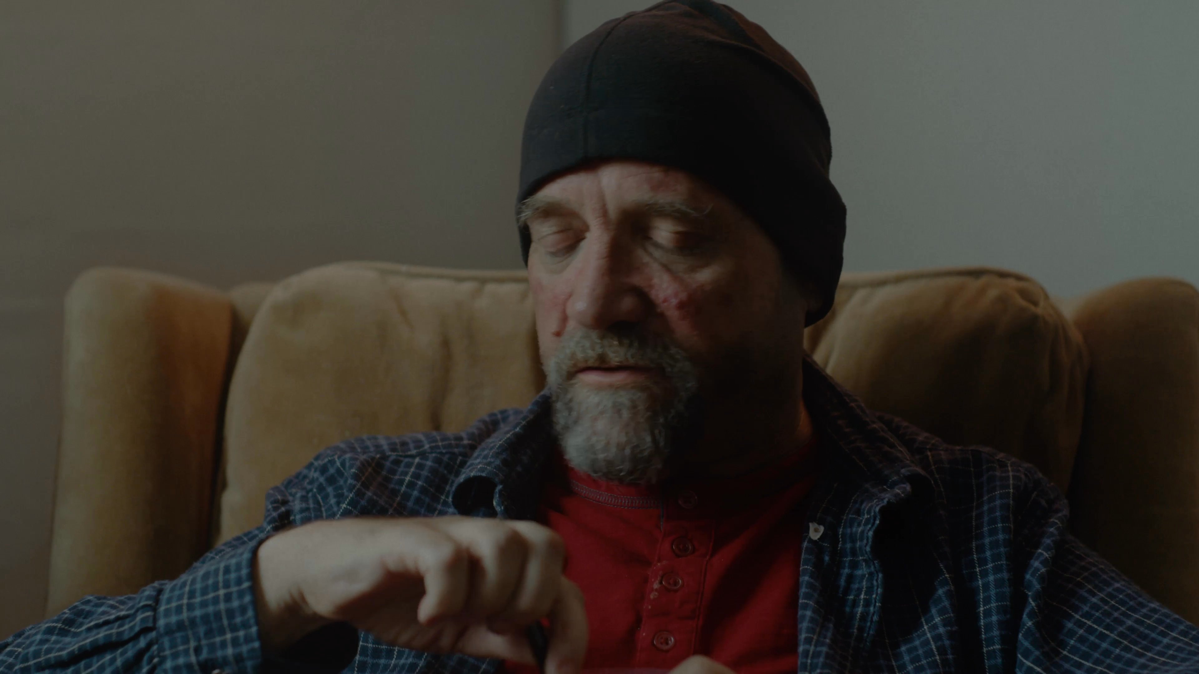 Two men engage in a calm conversation while seated in a domestic living room environment. The scene emphasizes their interaction as they exchange dialogue in an informal setting.